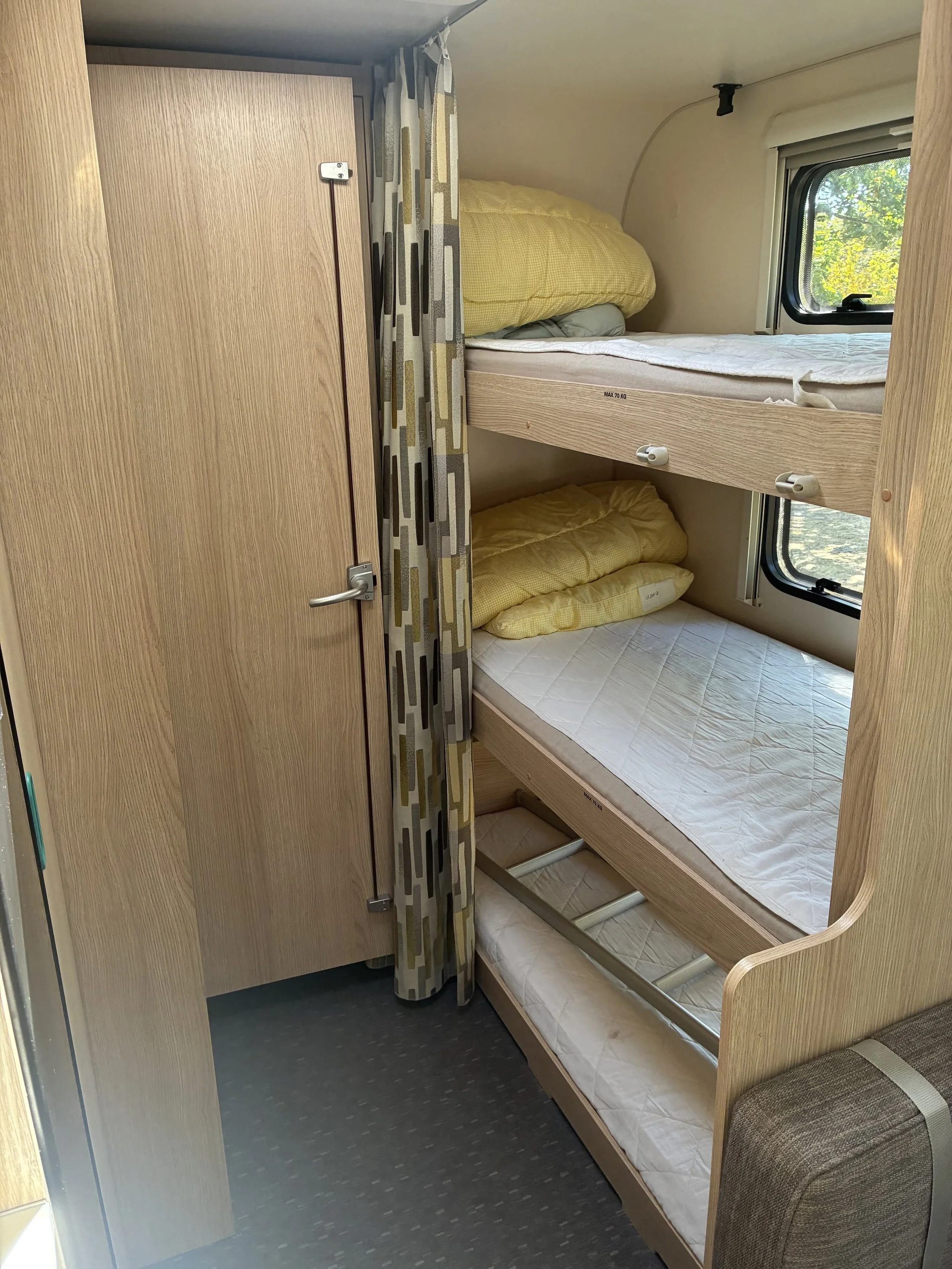 Indretningsbillede af en sovecabine med tre træk-senge, hvoraf den øverste har en pude, og der er en vinduesramme med udsigt til grønne blade.