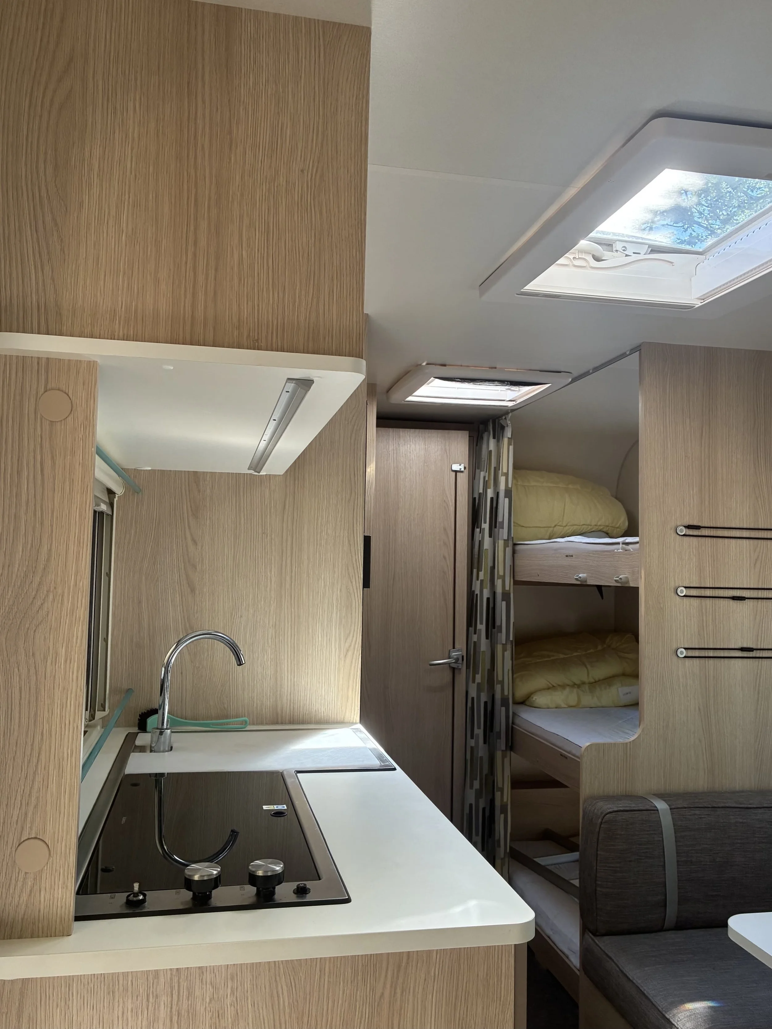 Indendørs billede af et lille køkken med kokeplade og vask, samt bunksenge med gule dyner og store ovenlys i en campingvogn.