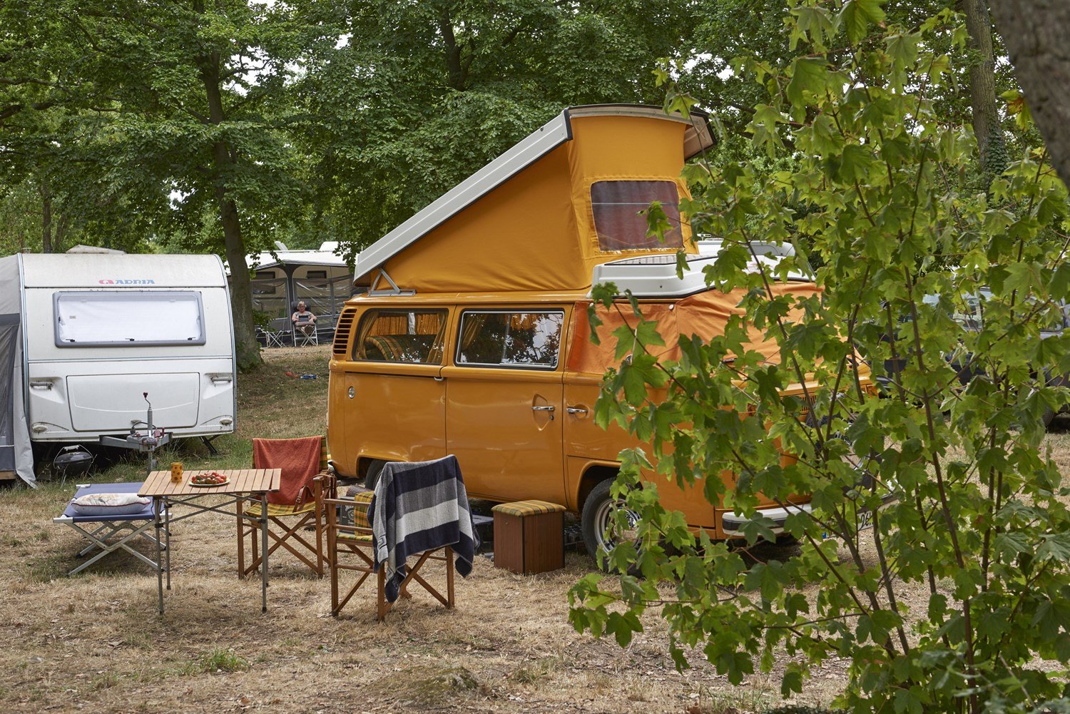 En orange campingvogn med tagtelt, omgivet af træer, med en opstillet udendørs spiseplads med borde, stole, og tæpper.