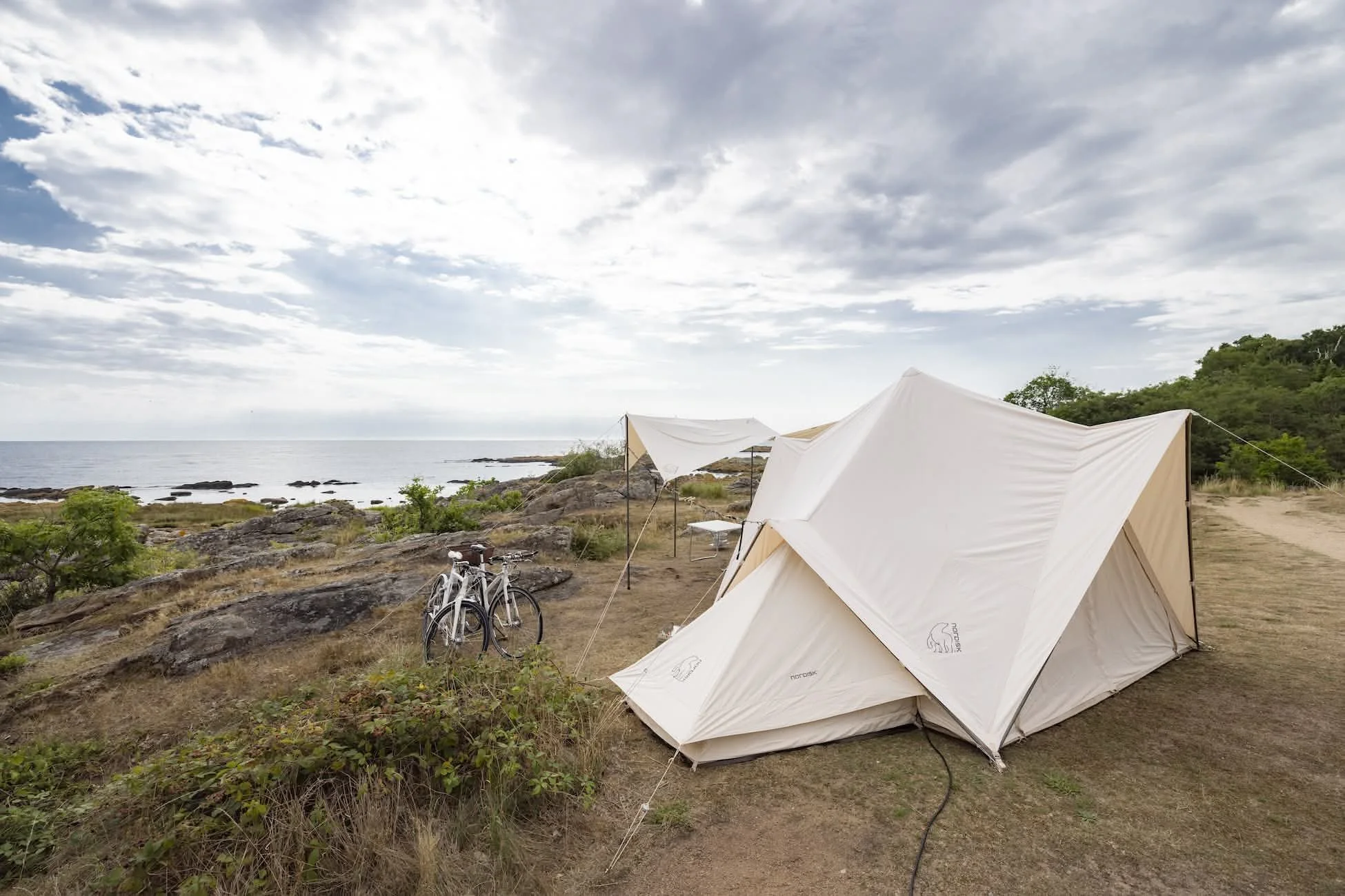 Camping på en strand med to hvide telte og cykler, hav og klipper i baggrunden, overskyet himmel.