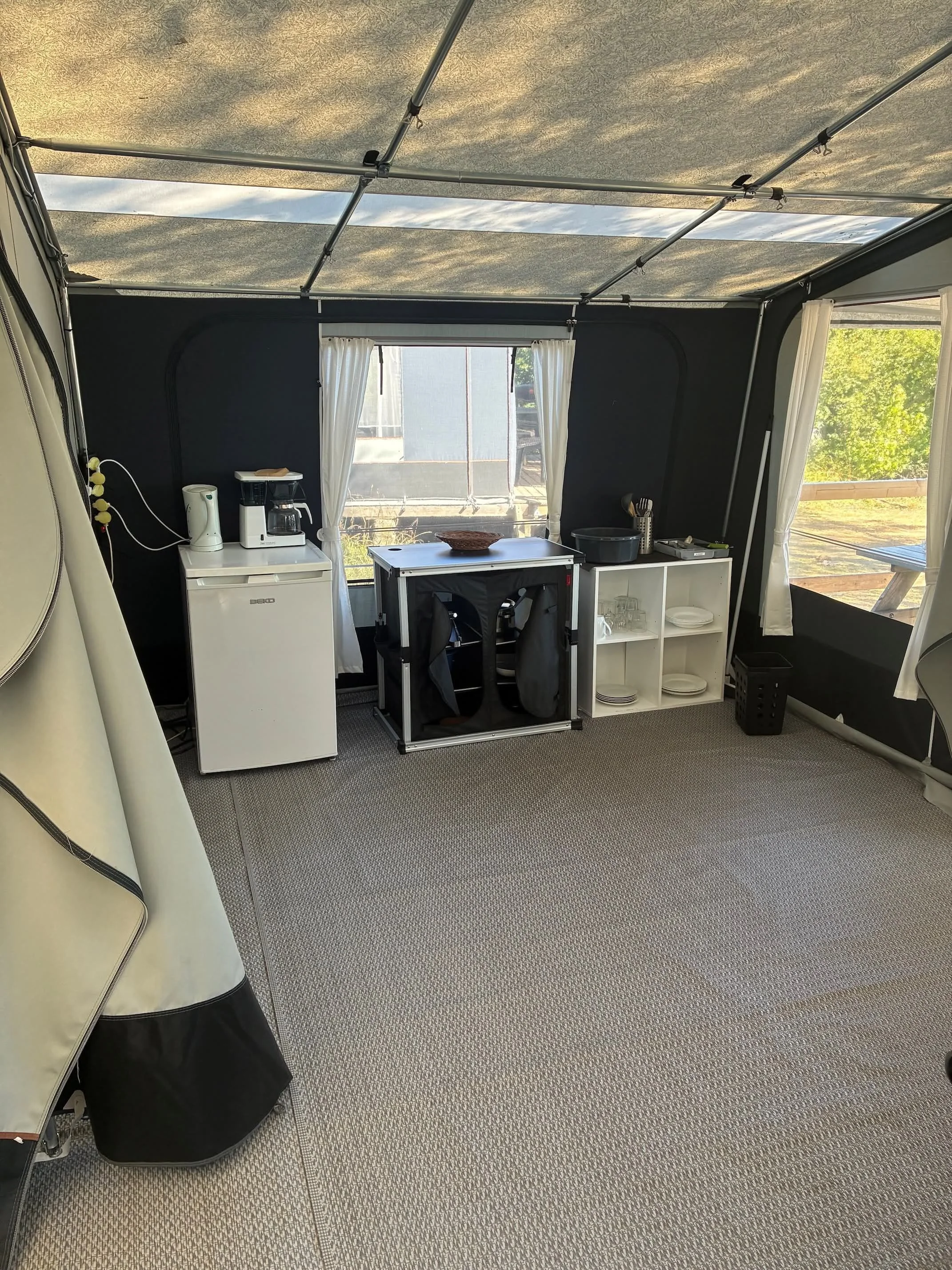 Indendørs campingvognsinteriør med kaffemaskine, minifryser, skab, tallerkener, glas og service.