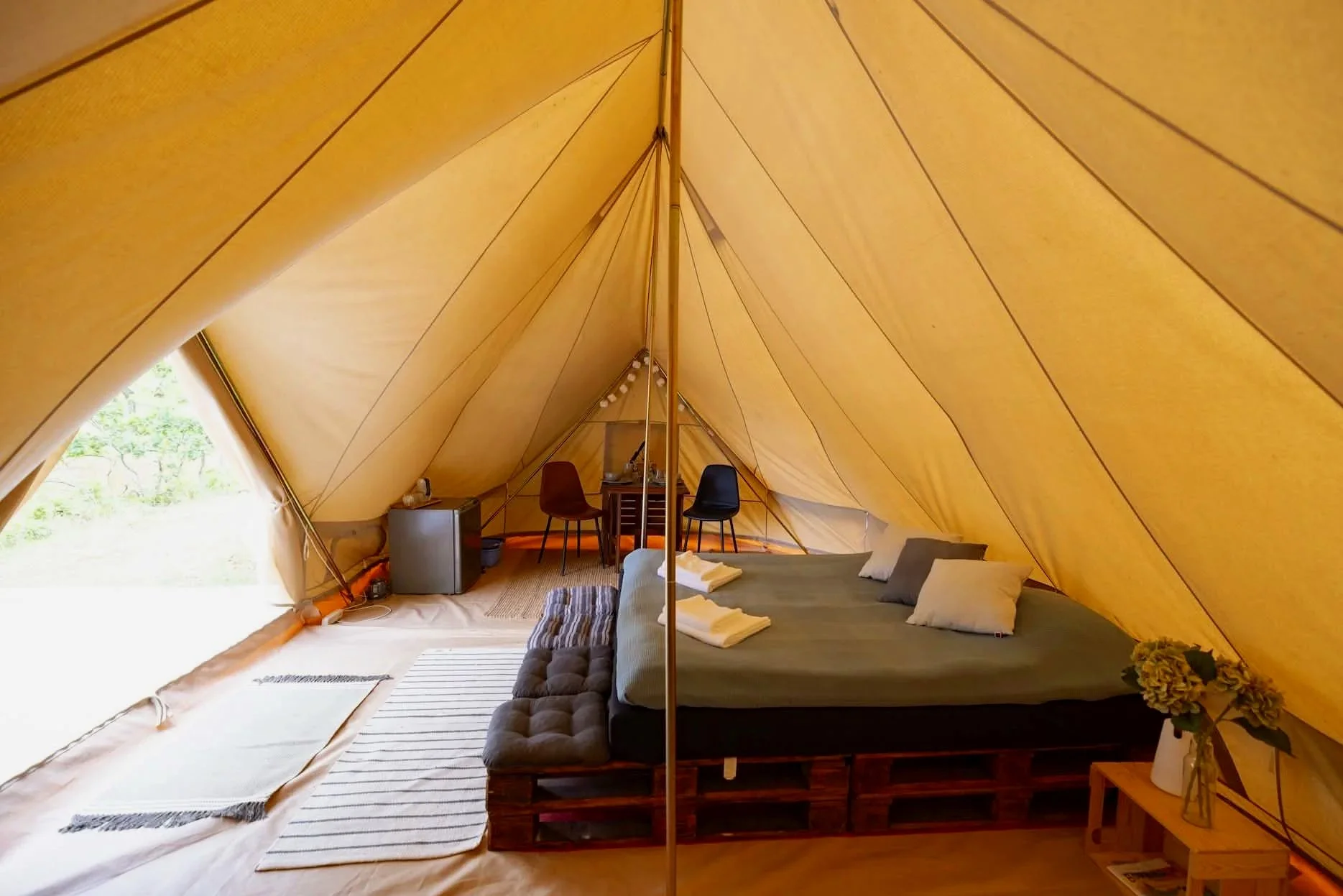 Indretning af en campingtelt med en stor seng, små tæpper, en plante på et lille bord, to spiseborde med stole, samt et lille køleskab.