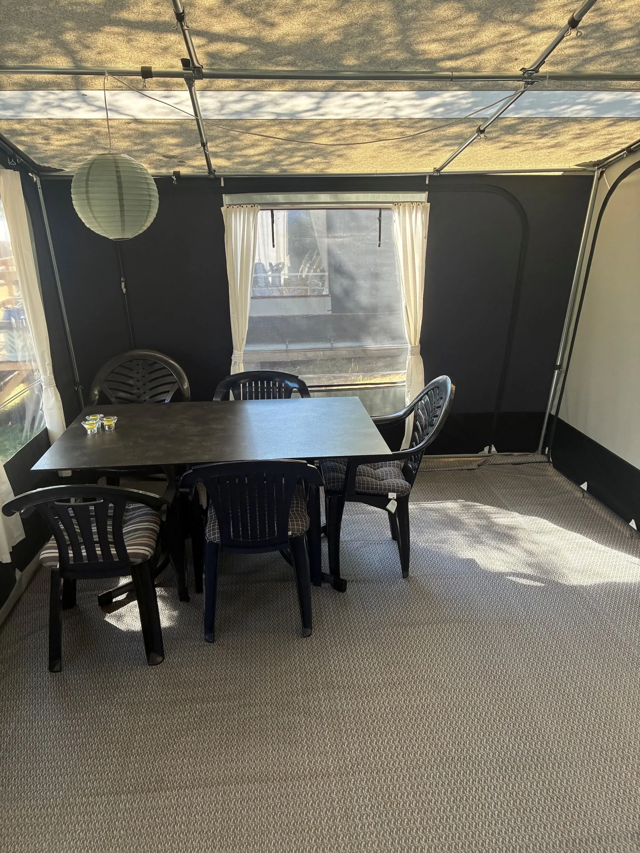 Indendørs terrasse med sort bord, seks sorte plastikstole, en hvid lampskærm, vinduer med hvide gardiner og tæppe på gulvet.