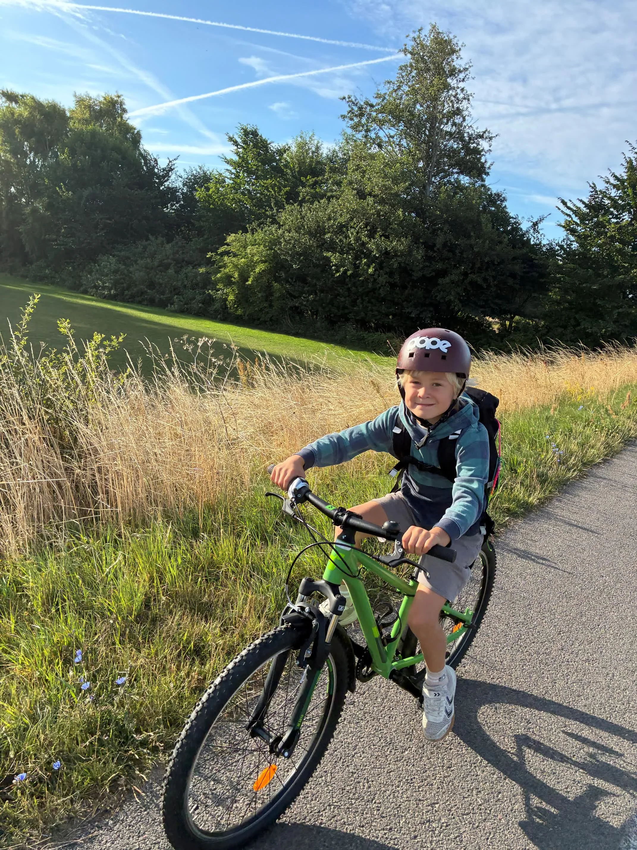 En dreng med hjelm cykler på en grusvej omgivet af græs og træer under en blå himmel.