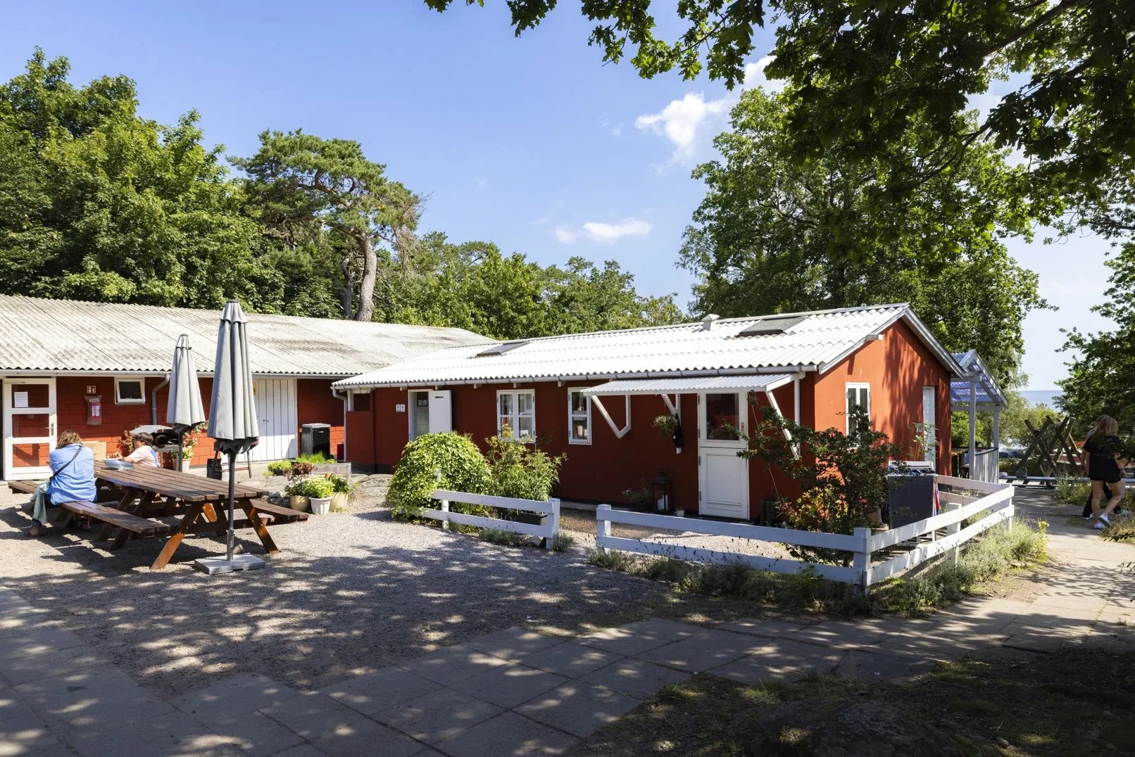 Rødt træhytte med hvid dør og vinduer, omgivet af grønne træer, med udendørs siddepladser og parasoller på en solrig dag.