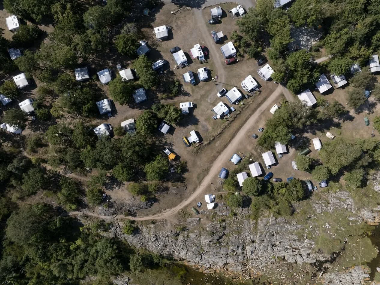 A drone view of a campsite with numerous tents and caravans surrounded by trees, with a dirt path running through the area.