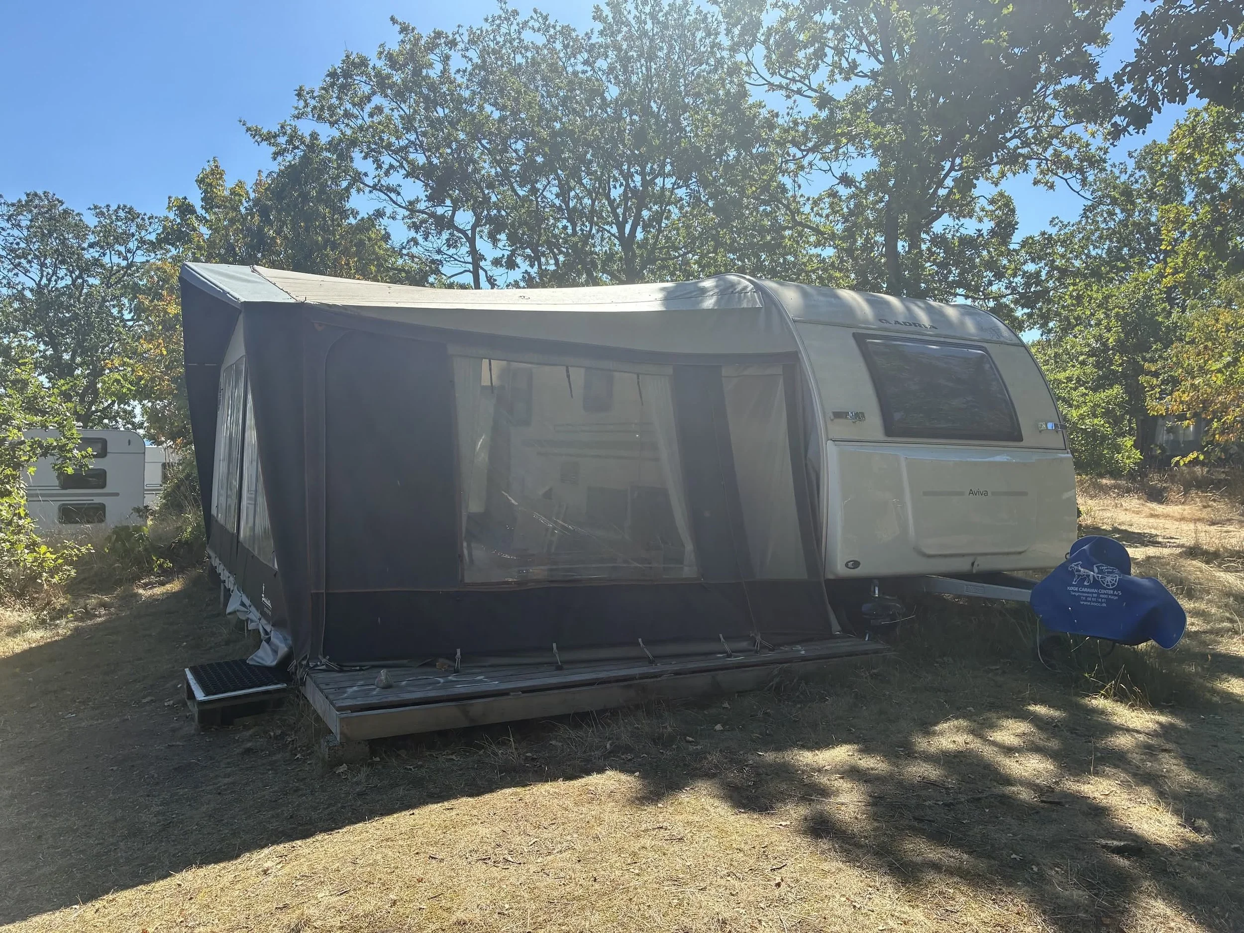 En campingvogn parkeret under træer med en markise udvidet, på en grus- og jordbund.