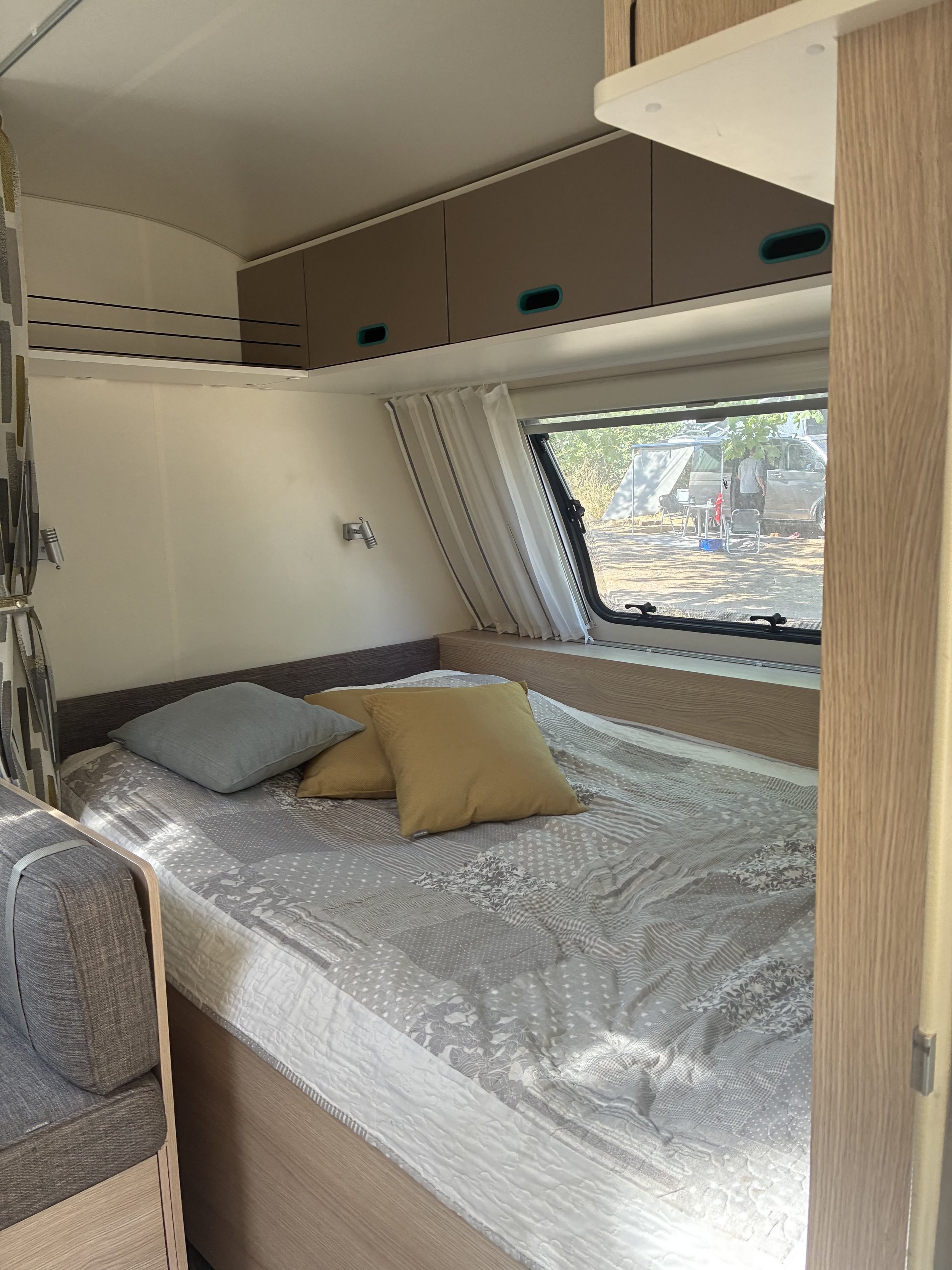 Indvendigt i en campingvogn med seng, puder, væglamper, garderobeskabe og et vindue med gardiner.