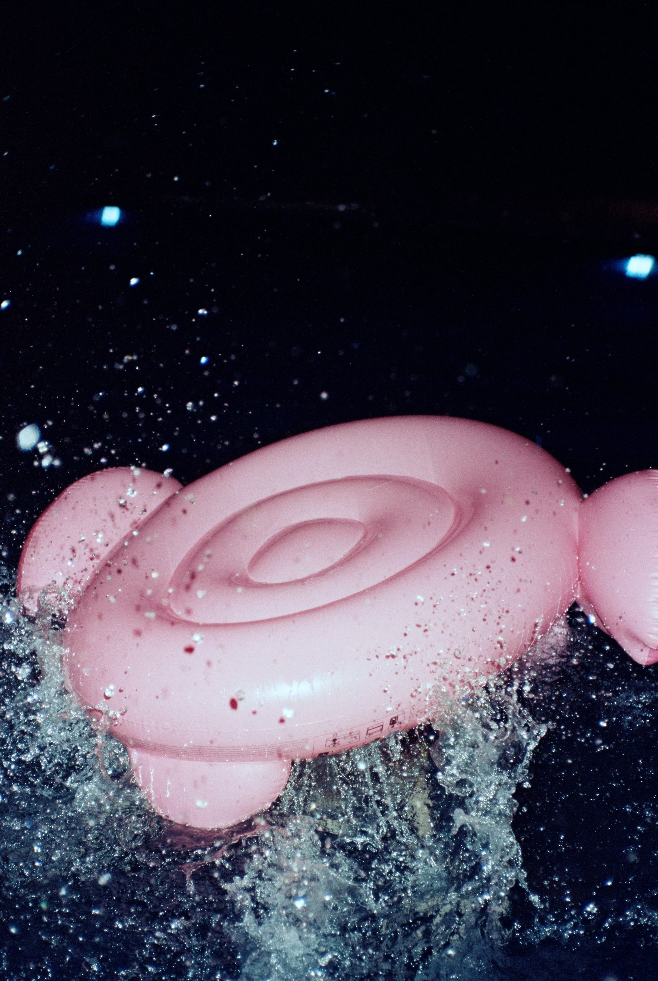 Pink inflatable pool float shaped like a donut with sprinkles, floating in water with splashes and bubbles around it.