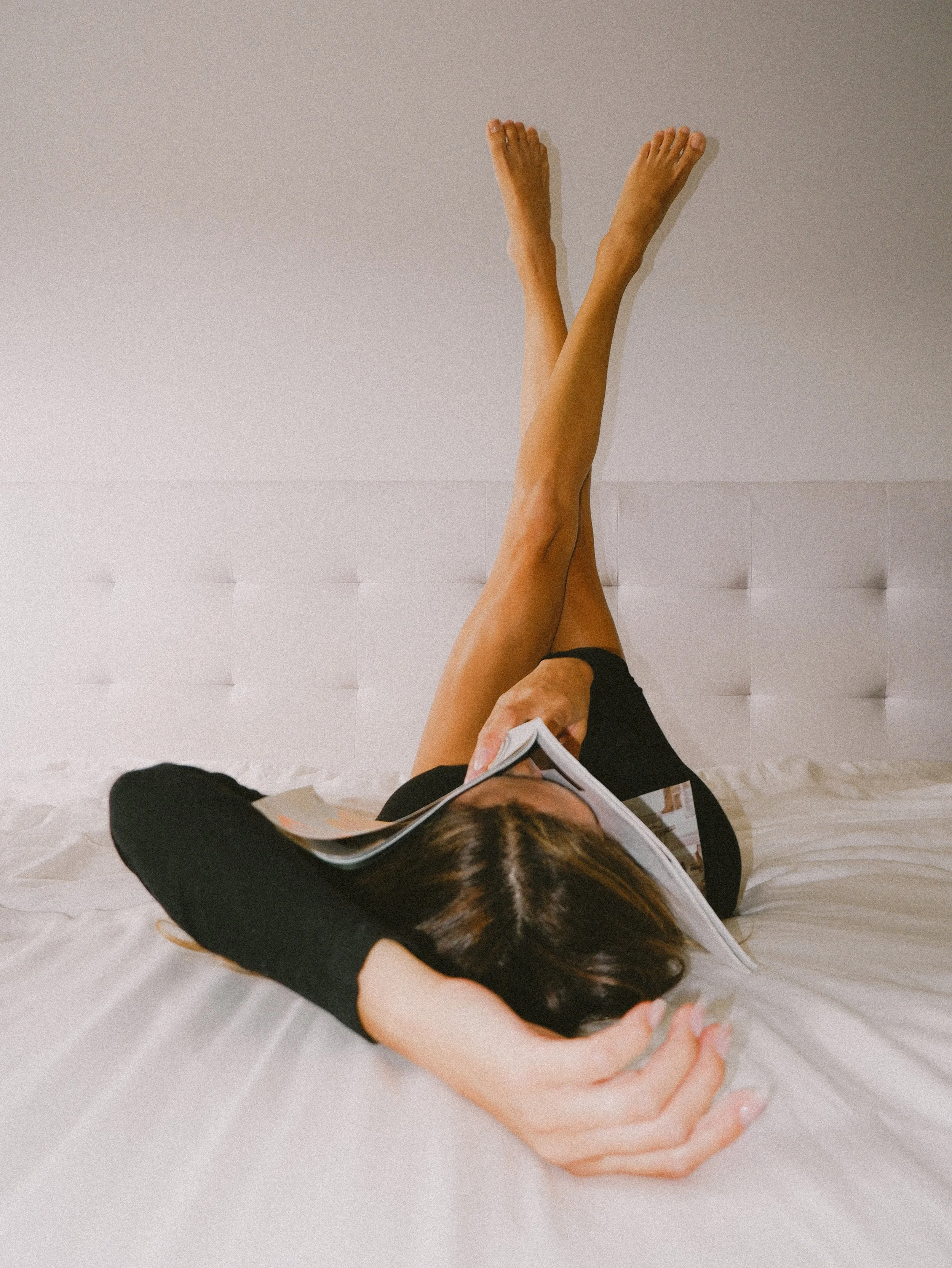 Woman lying on bed with legs propped against the wall, reading a book.