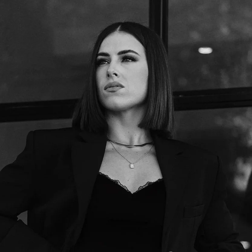 Black and white photo of a woman with shoulder-length dark hair, wearing a blazer and necklaces, looking thoughtfully to the side.