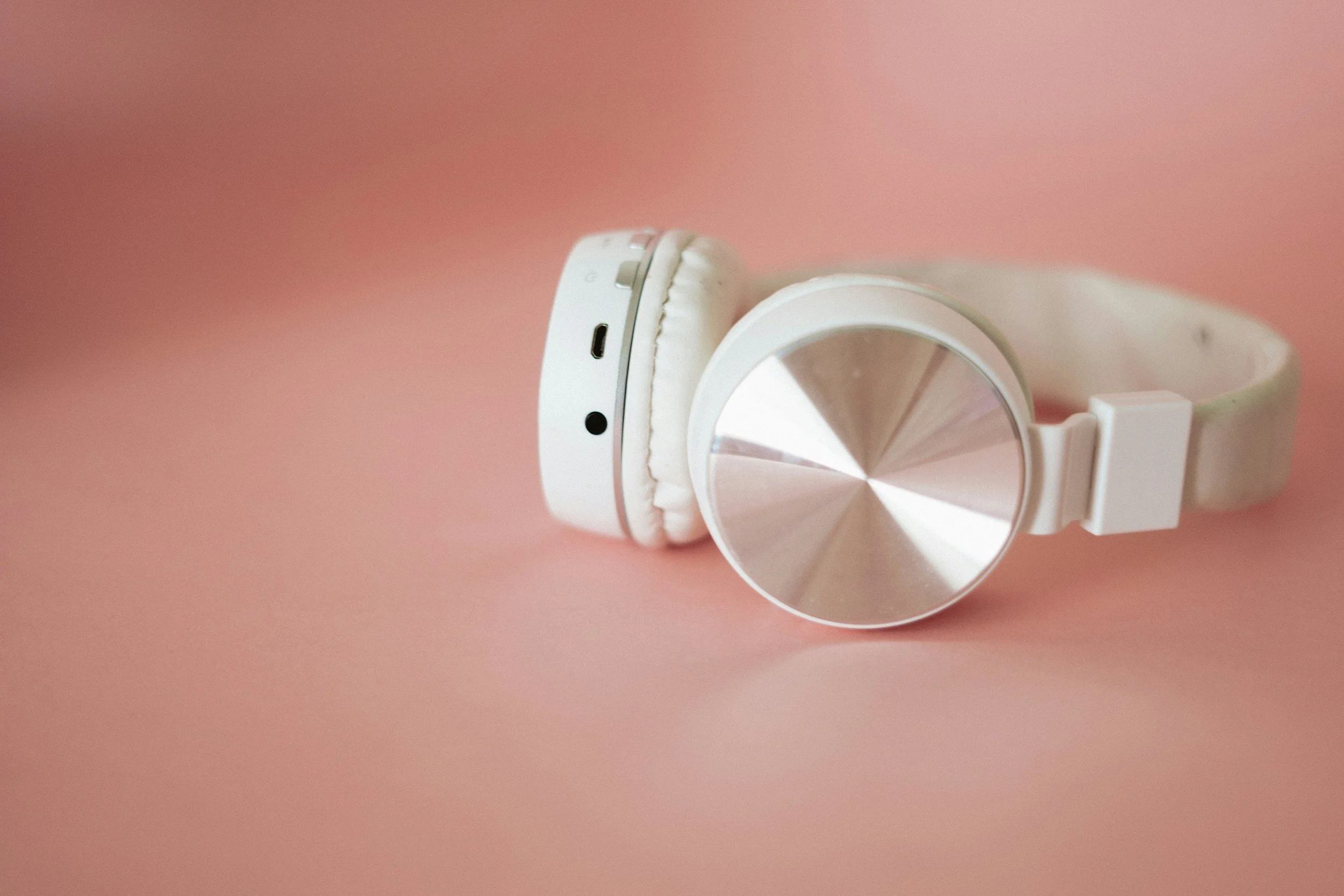 White over-ear headphones with a metallic circular design on the ear cup, resting on a pink surface.