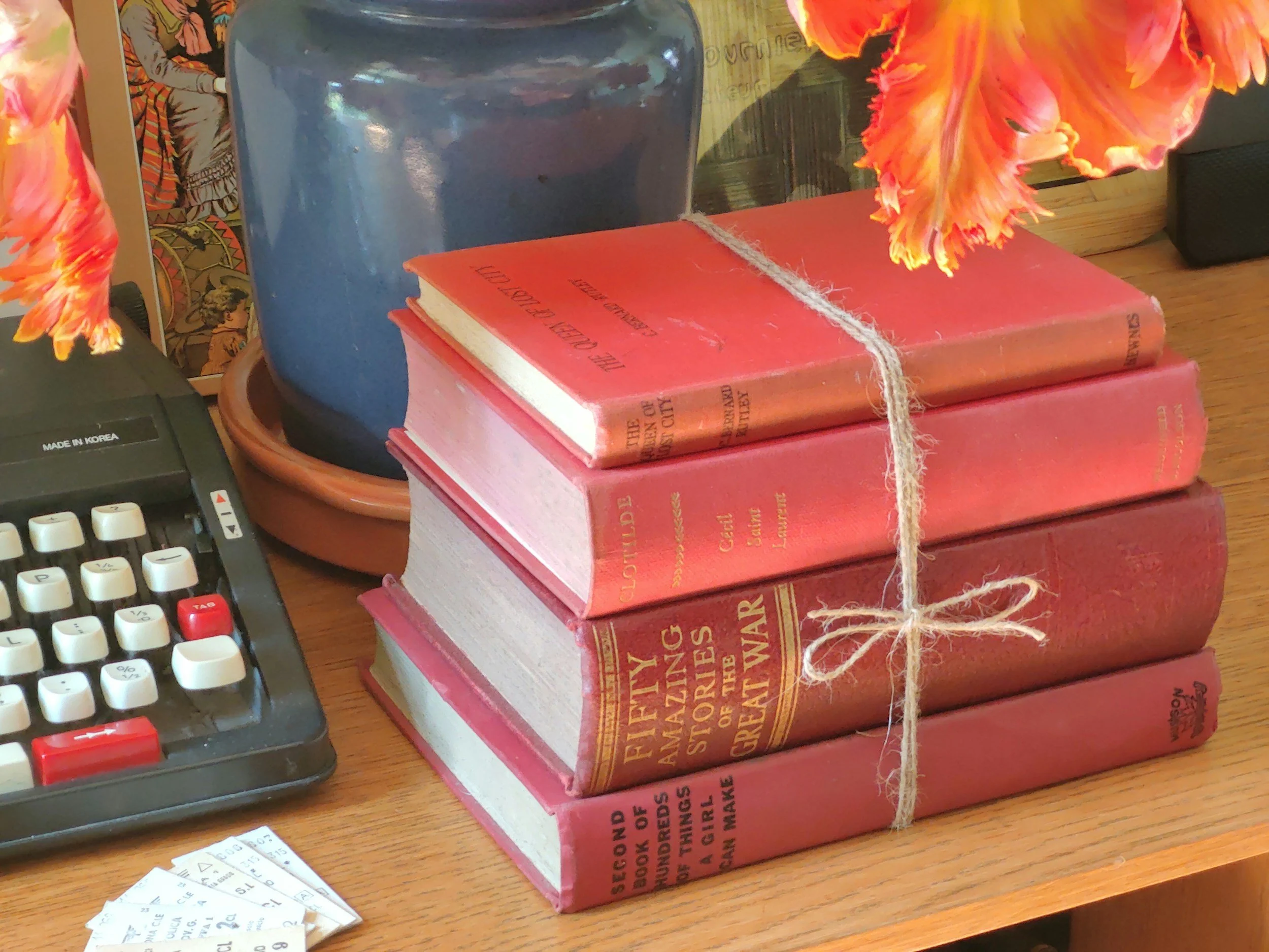 A stack of five red books tied with a string, placed on a wooden surface with a potted plant with orange pink flowers nearby.