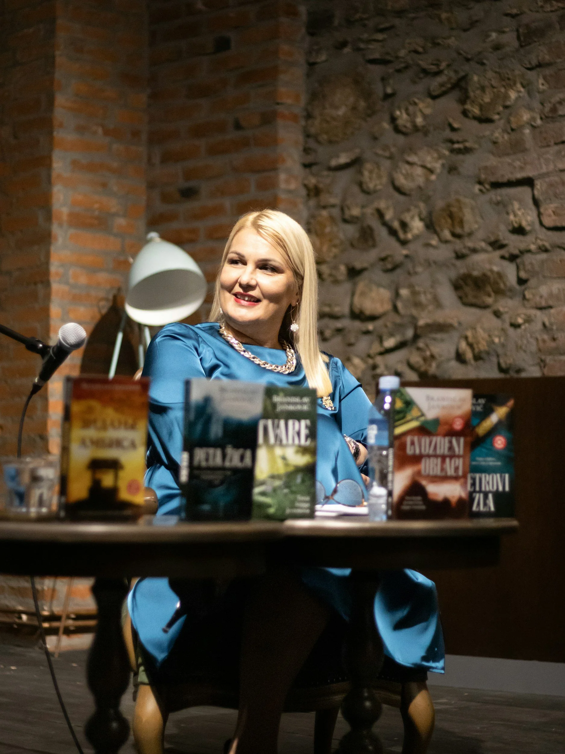 A woman sitting at a table with books, a water bottle, and a microphone in front of her, smiling, in a room with a brick and stone wall background.