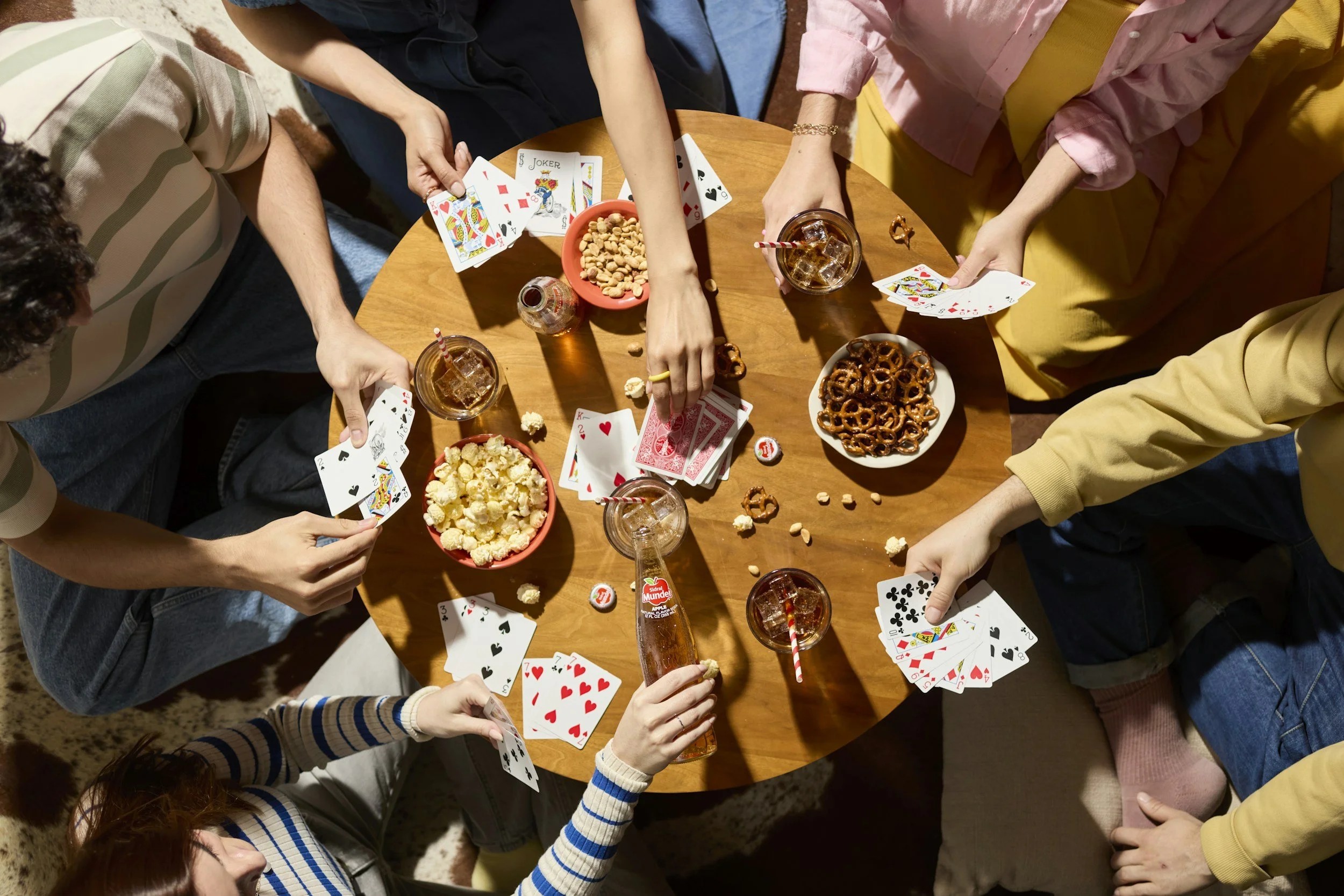 A group of people playing poker around a wooden table with snacks and drinks, viewed from above.