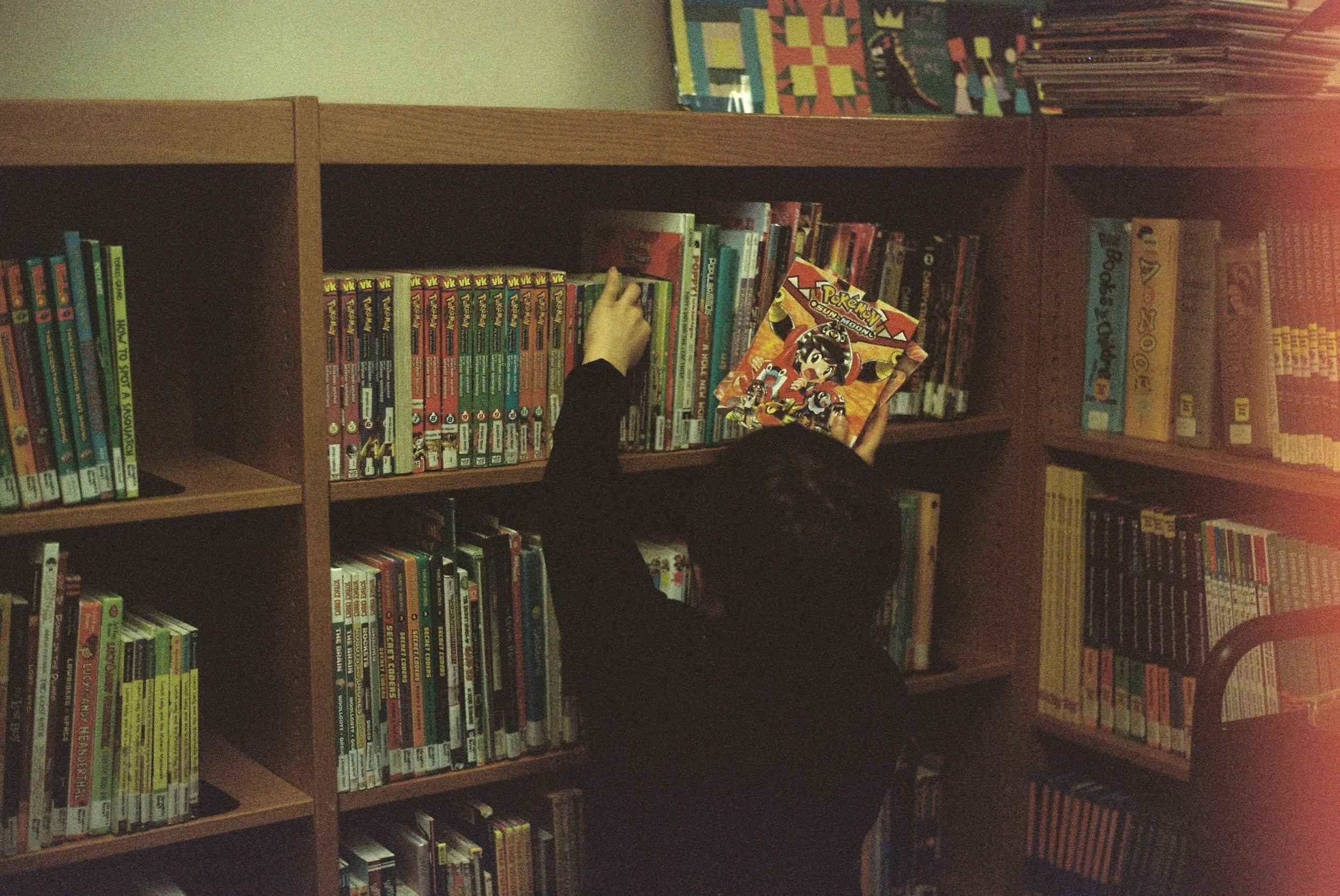 A person reaching for a comic book on a bookshelf in a library, surrounded by other books and comics.