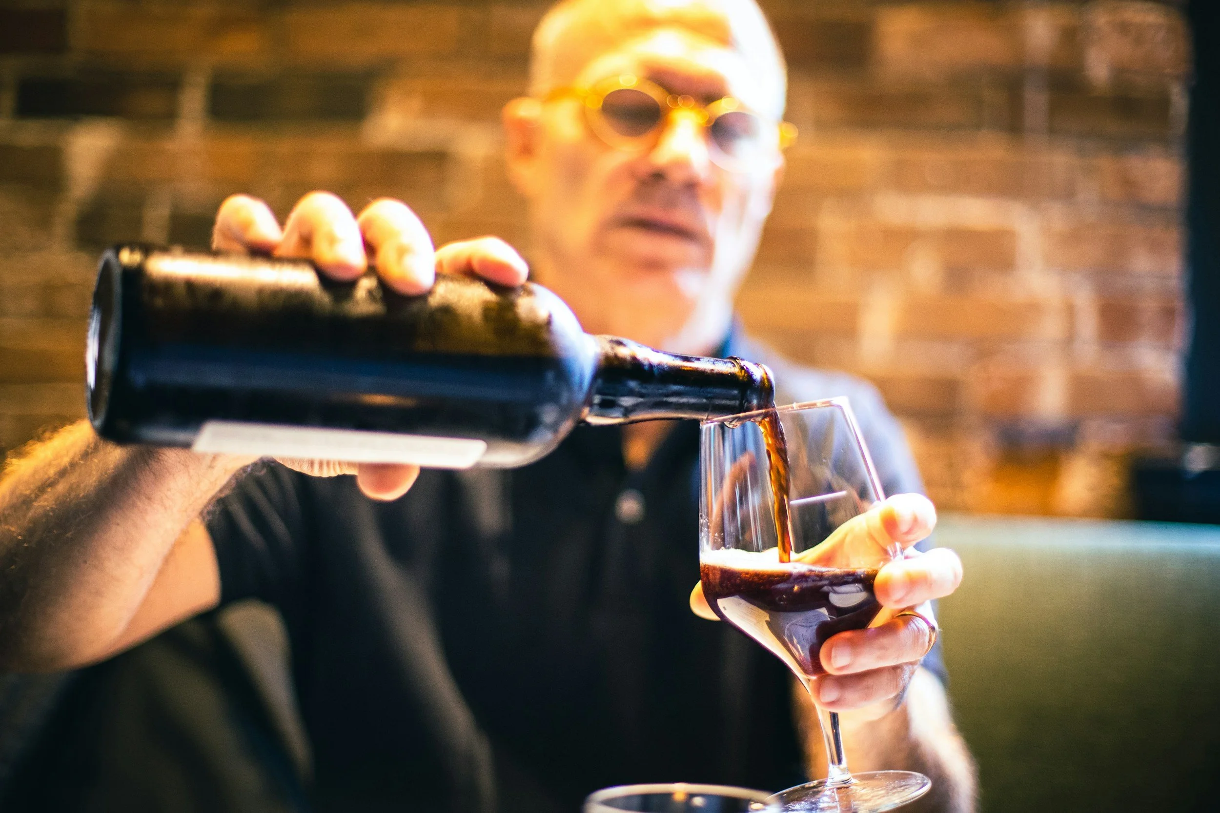 A man with glasses pours red wine into a wine glass at a winery.