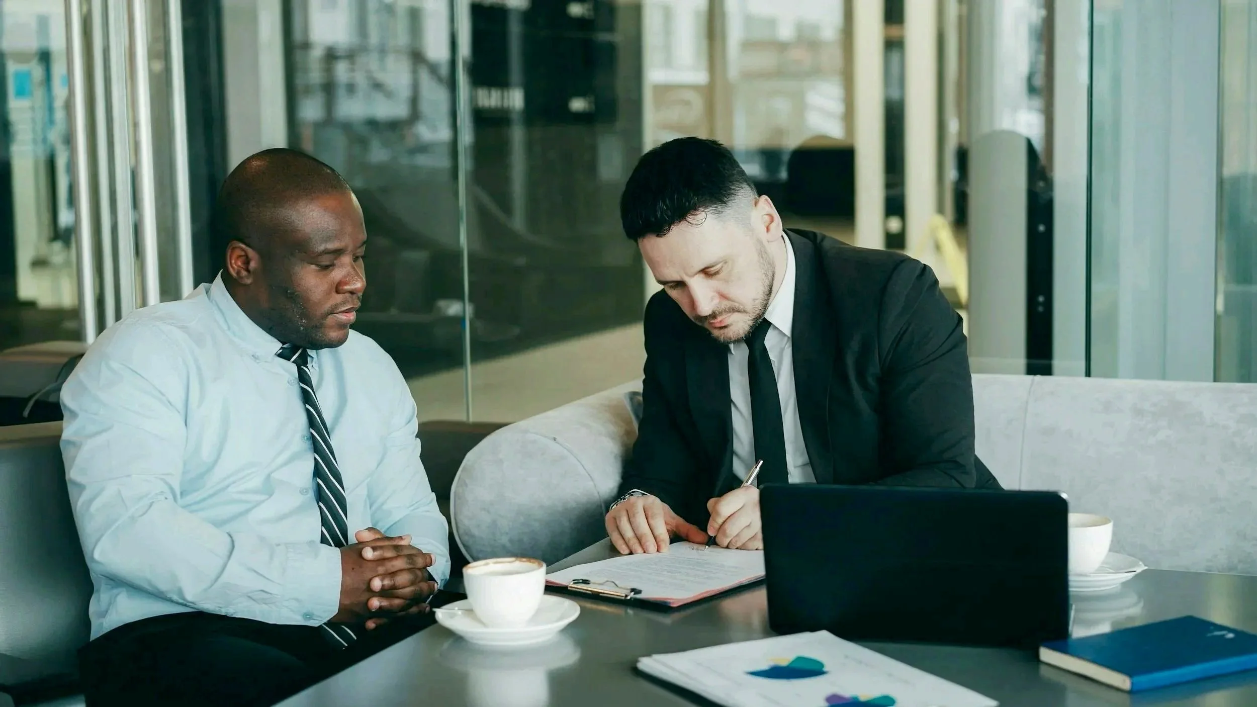 Two businessmen having a serious discussion in a modern office space, with a laptop, documents, and coffee cups on the table.