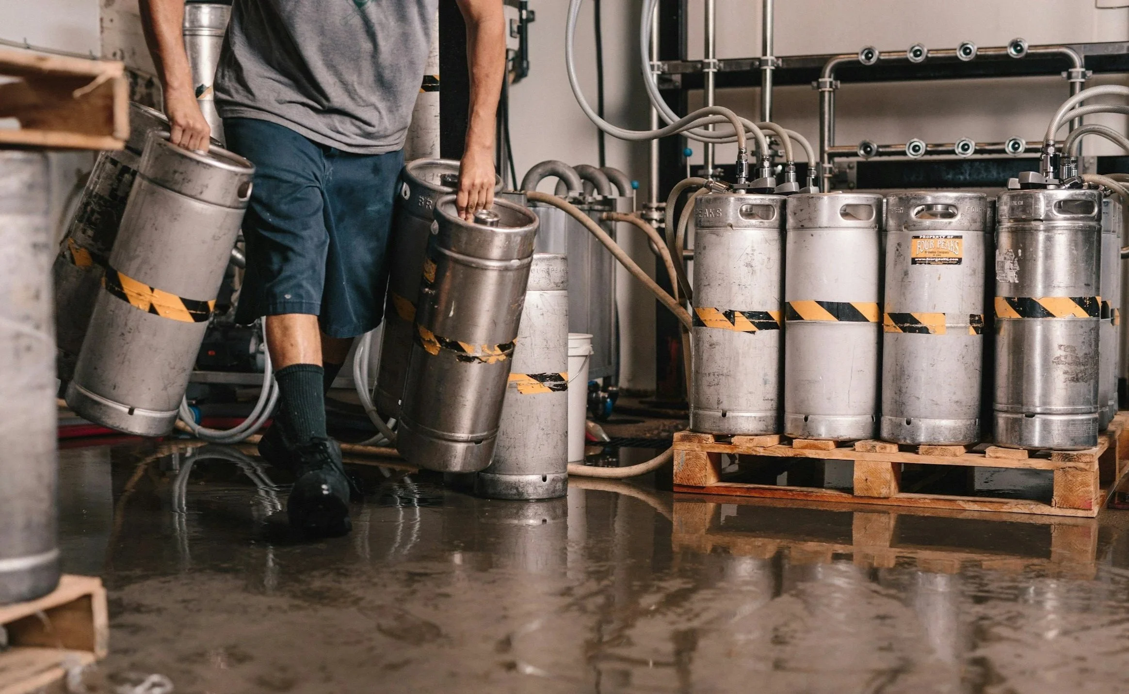A person is handling stainless steel tanks in a brewery or industrial facility, with pipes connected to the tanks and a wooden pallet in the background.