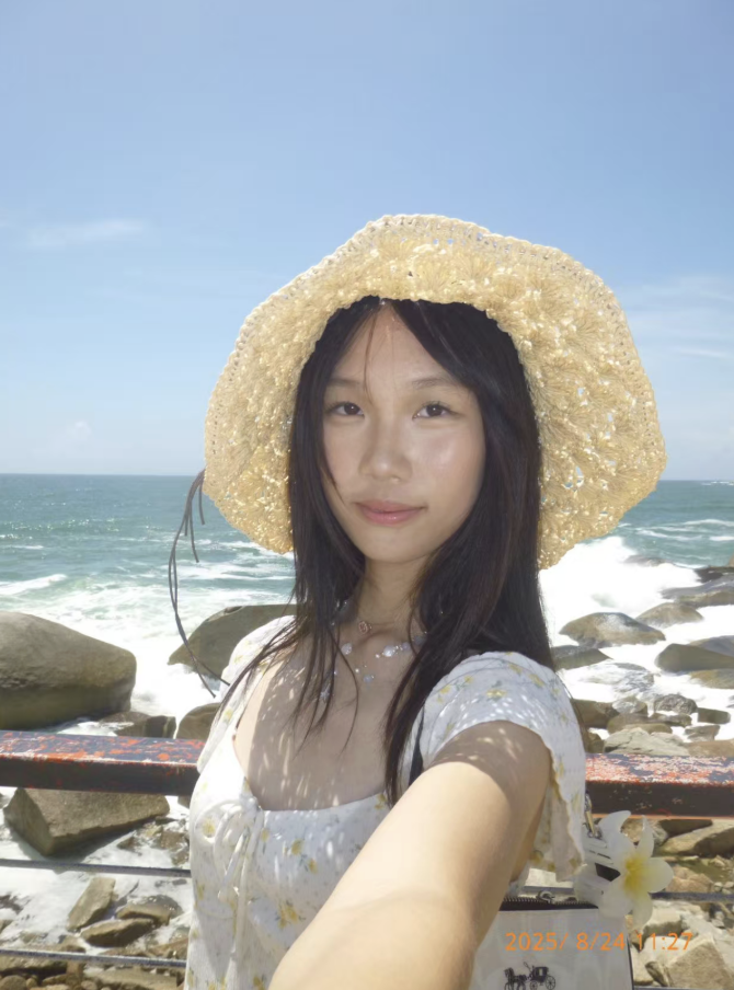 Young woman taking a selfie on a rocky beach with the ocean and blue sky in the background, wearing a wide straw hat and a white floral dress.