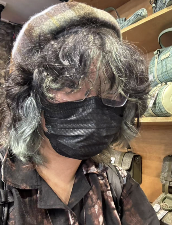 A person with curly hair wearing a black face mask, glasses, a hat, and a patterned shirt in a store aisle with bags and accessories on the shelves.