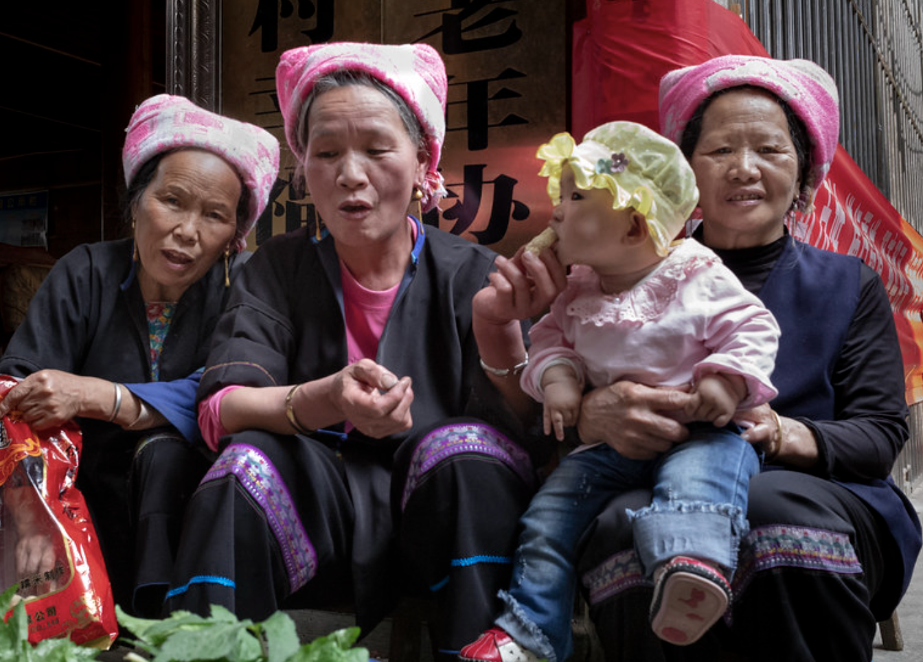 Four elderly women and one young child sitting together, all wearing pink towels on their heads, in front of a wooden background with Chinese characters.