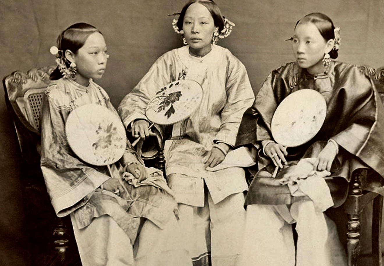 Three young Asian girls sitting on a vintage couch, dressed in traditional attire, holding fans and small brushes, in a black and white photograph.