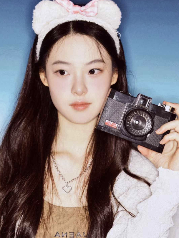 A young woman with long dark hair wearing a white fluffy headband with bear ears and a pink bow, holding a vintage camera to her face against a blue background.