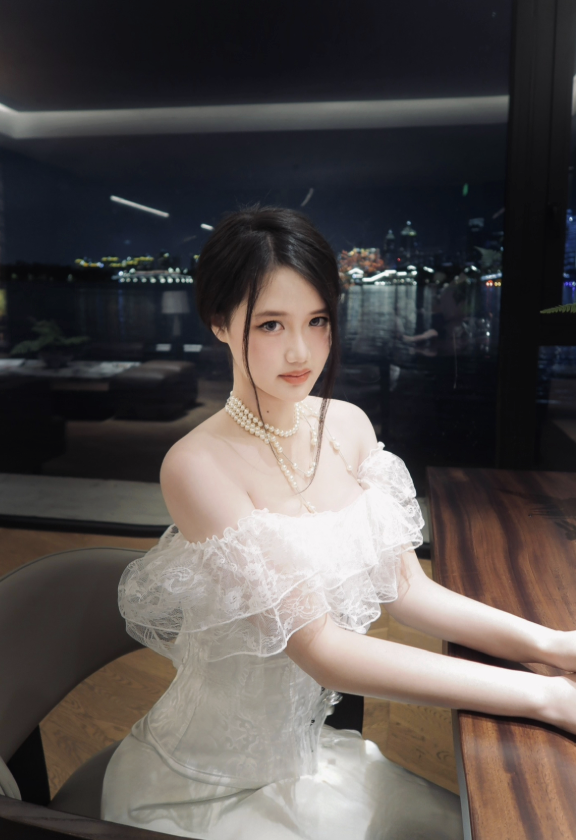 Young woman in an elegant white off-shoulder dress with lace ruffles, pearl necklace, and dark hair styled in a short bob, sitting at a wooden table by a window with a city skyline and river view at night in the background.