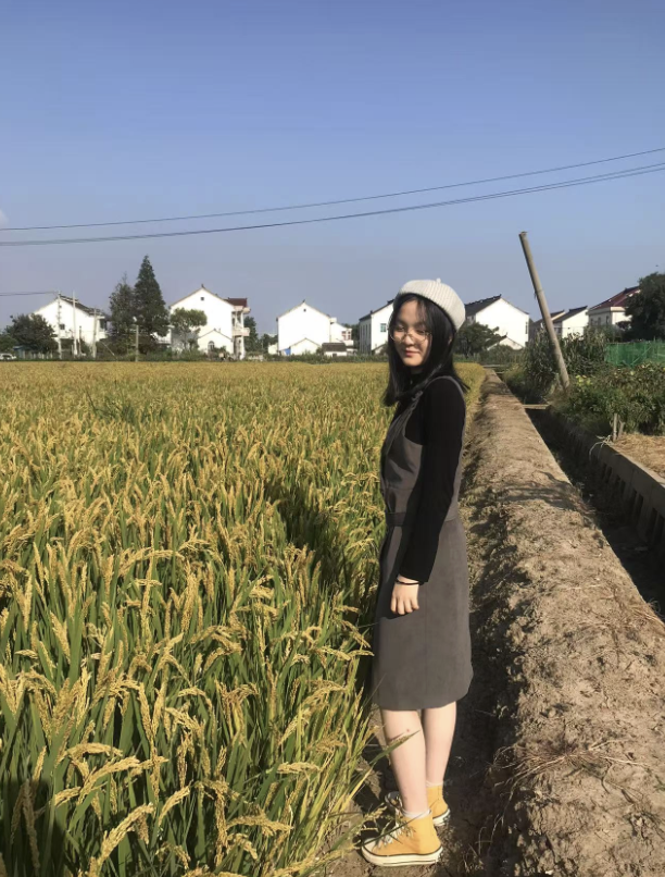 A young woman in a gray dress, black jacket, and white beanie hat standing on a narrow dirt path beside a golden rice field, with a row of white houses in the background under a clear blue sky.