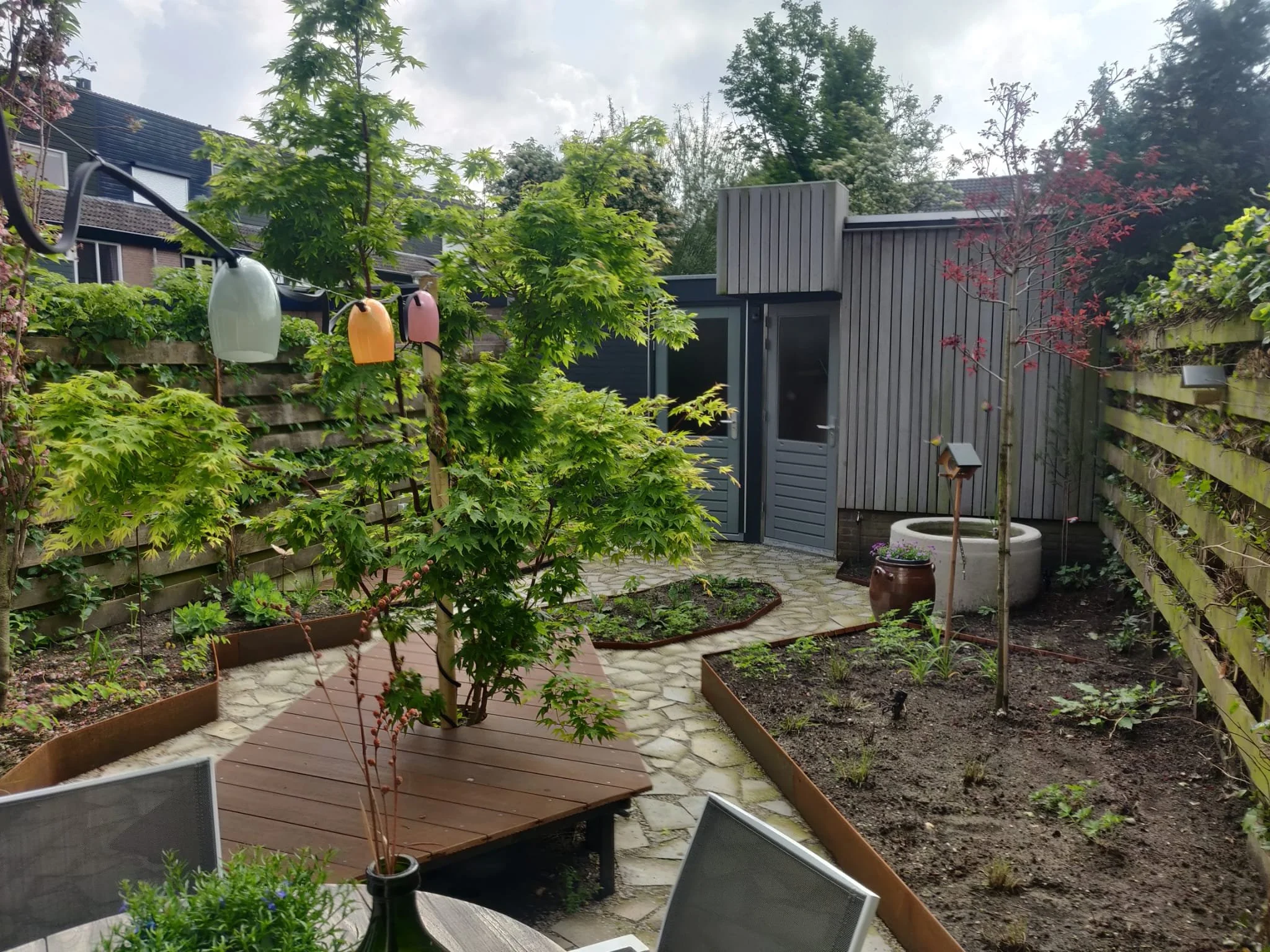 A backyard garden with a wooden deck, a central tree, string lights, and a small shed, surrounded by a wooden fence, with flowering plants and a potted plant with purple flowers.
