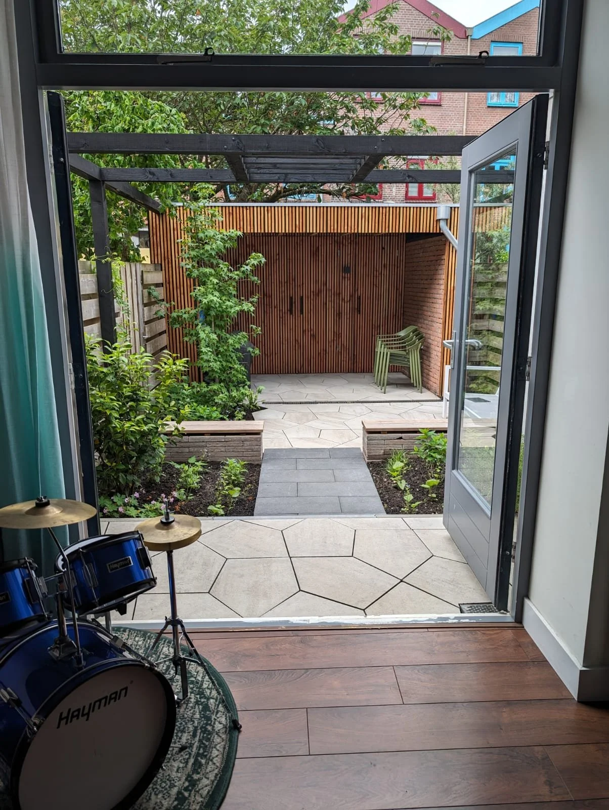 View from inside a house looking out through an open door onto a small garden or patio area with plants, paving stones, and stacked chairs against a wooden fence with a trellis and neighboring houses visible in the background.