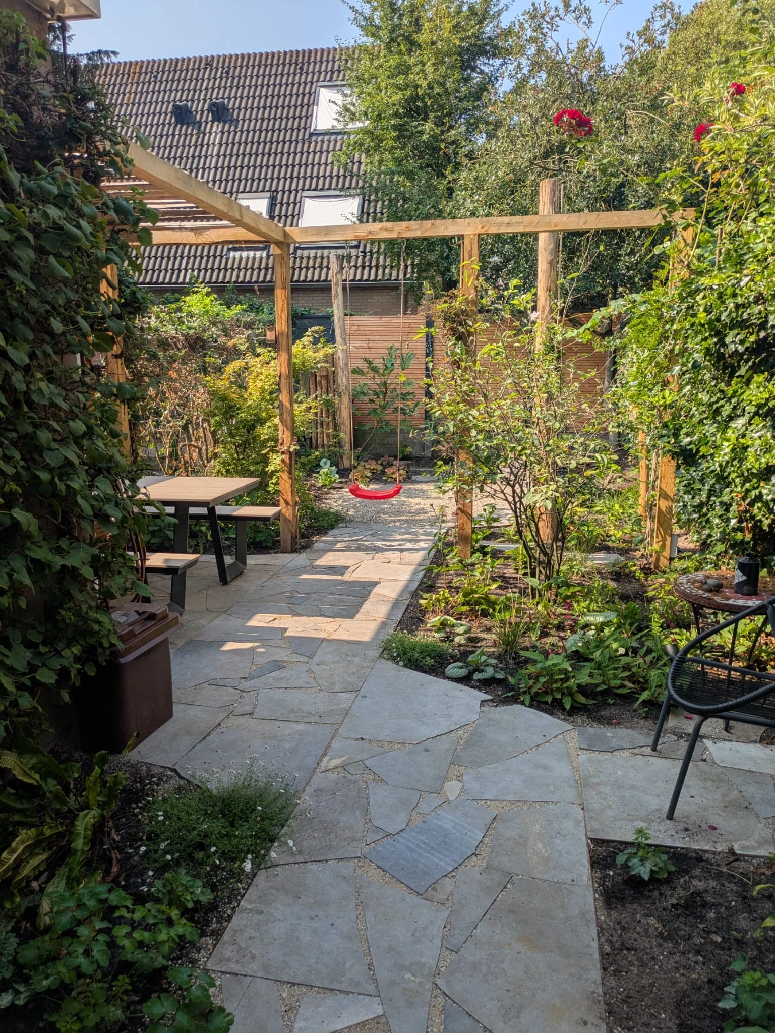 A backyard garden with a stone pathway, surrounded by lush green plants and trees. A swing hangs from a wooden frame, and there is outdoor seating including a table with benches and a black chair. The background shows a house with a tiled roof and skylights.