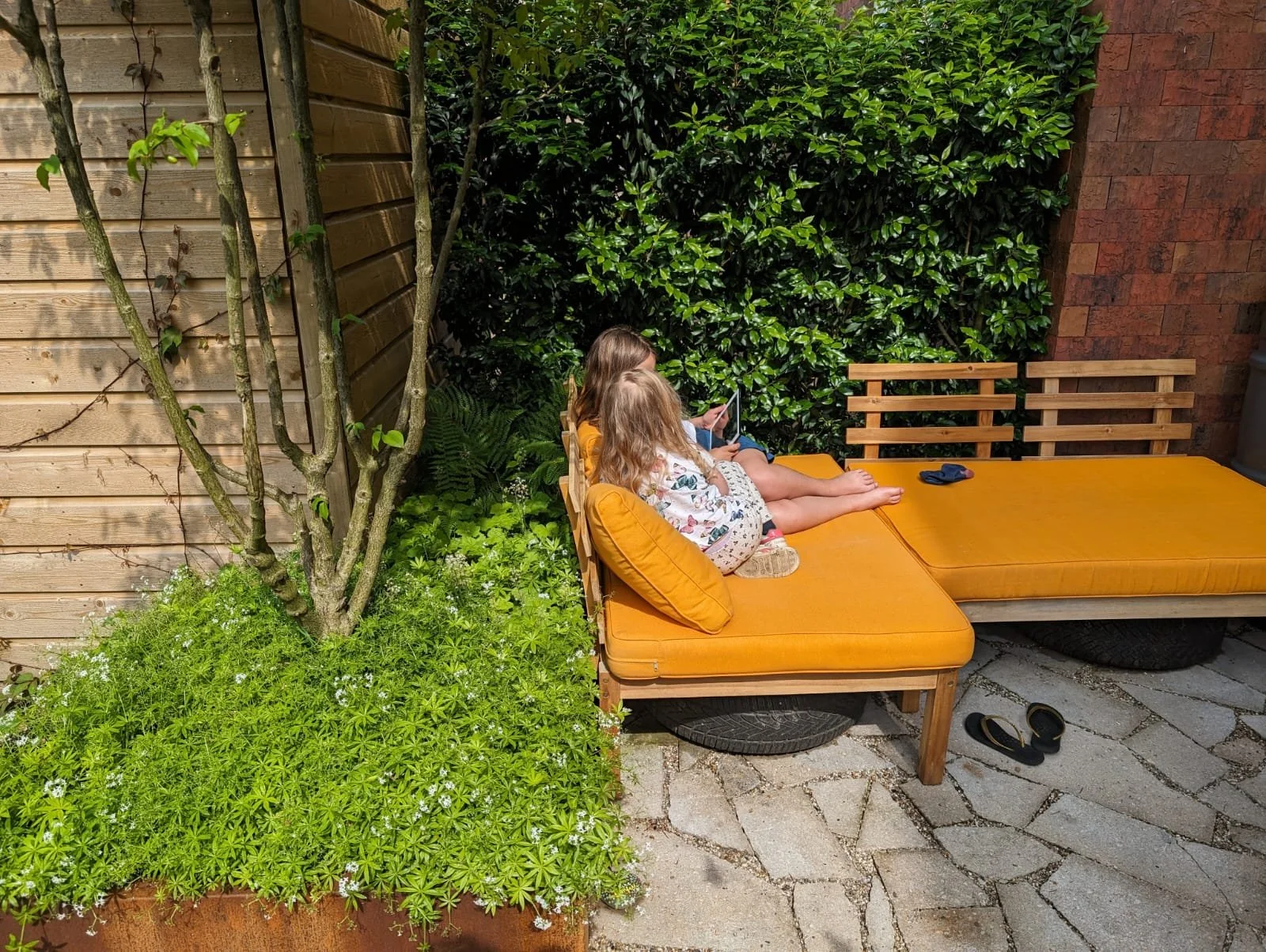 Two children with long hair sitting on orange cushions on a wooden outdoor lounge, with a tray of shoes on the ground, in a garden patio with plants, trees, a wooden fence, and a brick wall.