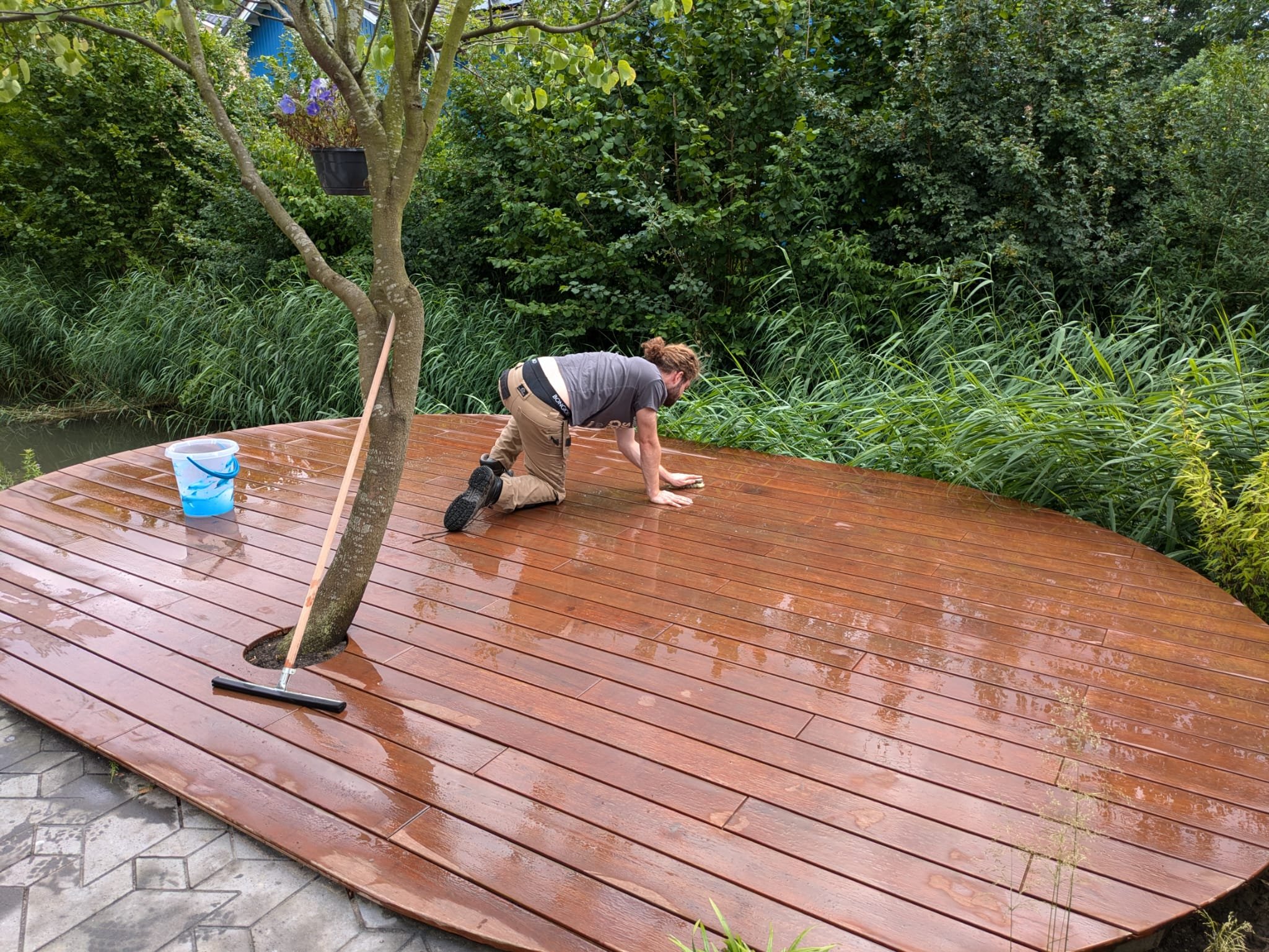 A man is cleaning a freshly installed wooden deck near a pond or lake, surrounded by lush green plants and trees. The man is kneeling on the deck, using a cloth or sponge, with a bucket nearby and a squeegee leaning against a tree.