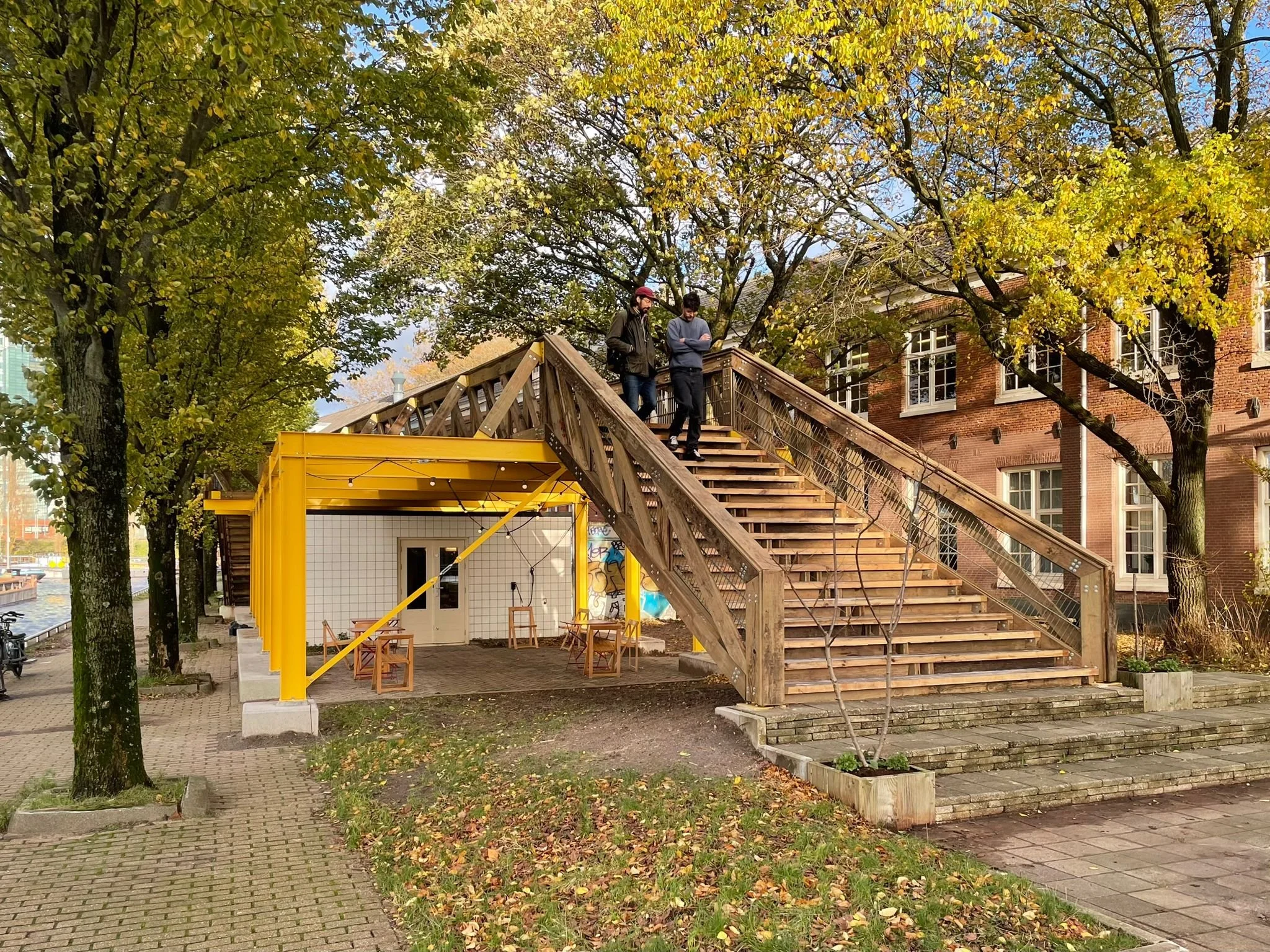 People walking down wooden stairs attached to yellow and wooden structures in an outdoor urban park with trees and historic brick buildings.