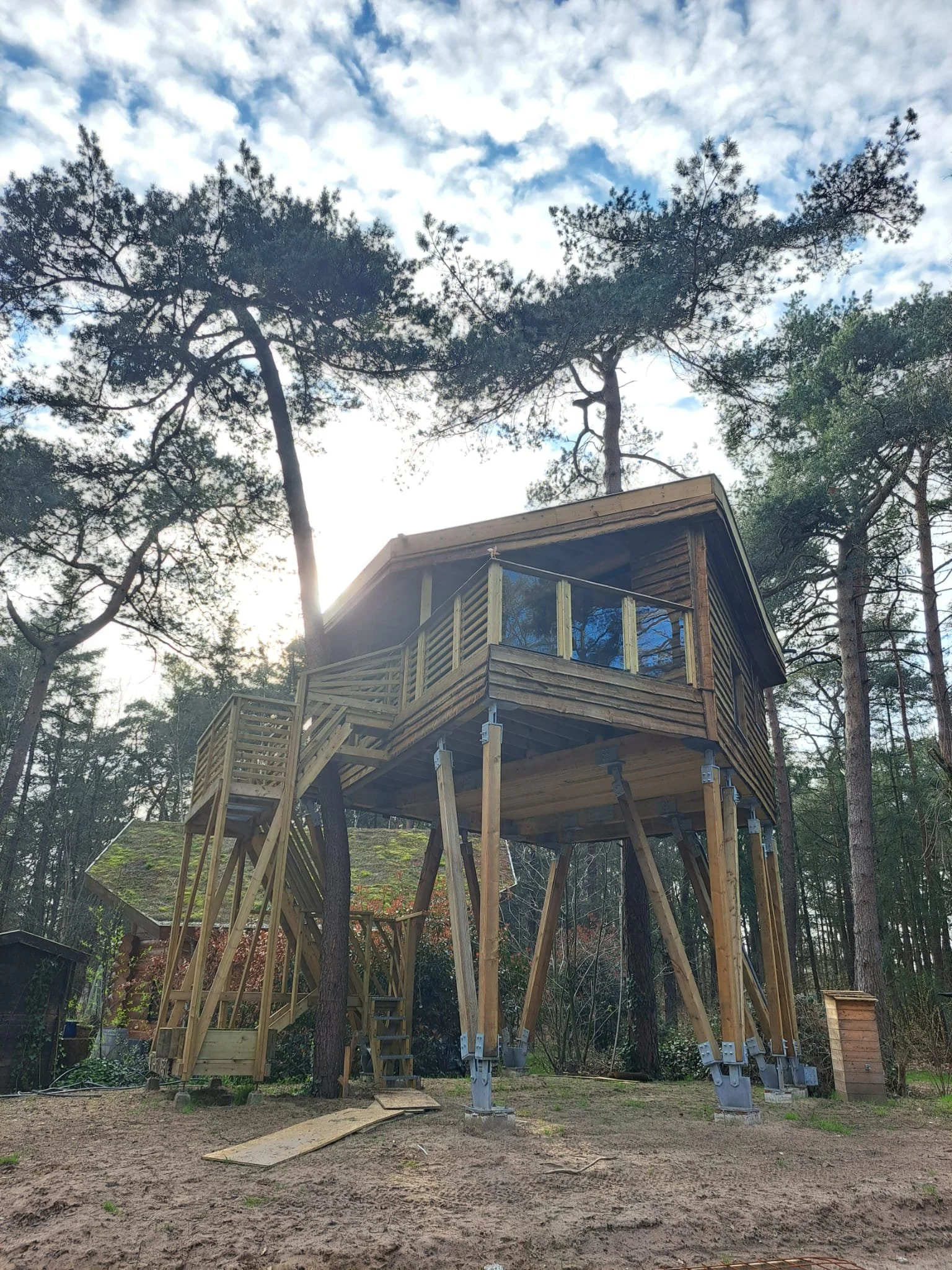 A wooden treehouse with a deck and stairway, elevated on stilts in a wooded area with trees and a cloudy sky.