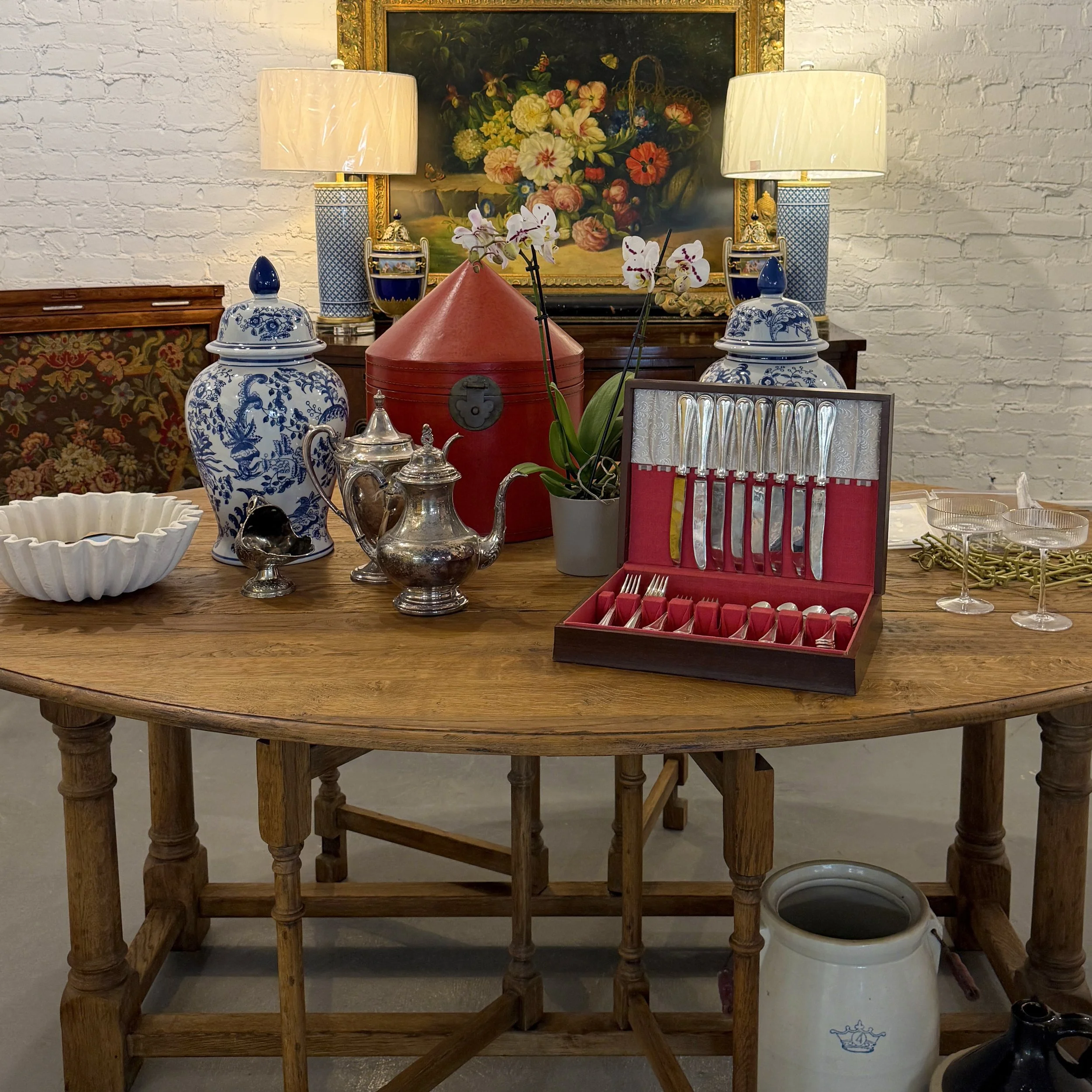 An antique wooden dining table with silverware, tea set, blue and white porcelain vases, a red box, orchid plant, and a set of knives in a case, with lamps and a framed floral painting in the background.