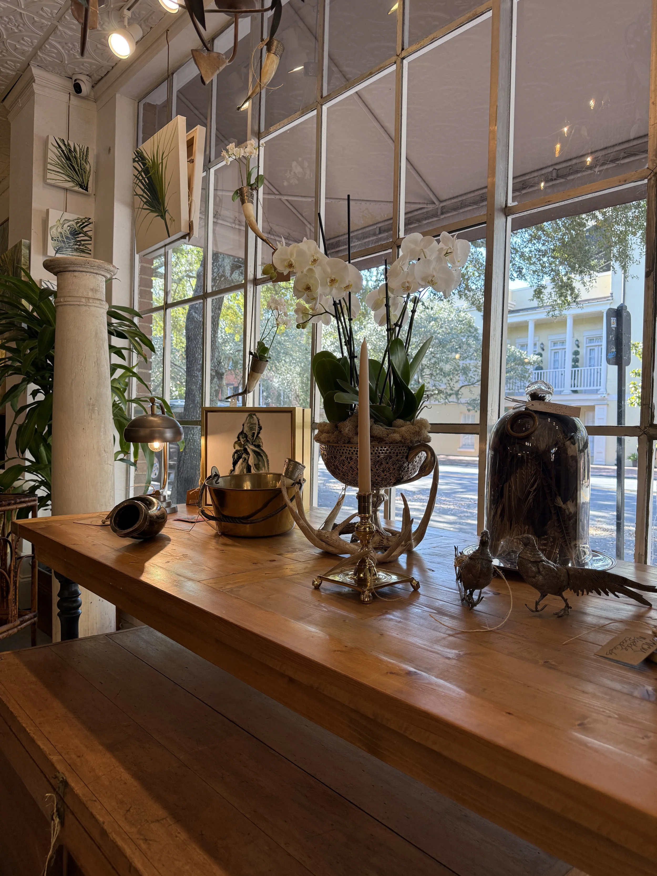 Decorative display on a wooden table featuring a potted white orchid, metal bird sculptures, a candlestick holder, a glass jar, and small framed artwork, with large windows in the background revealing trees and buildings outside.
