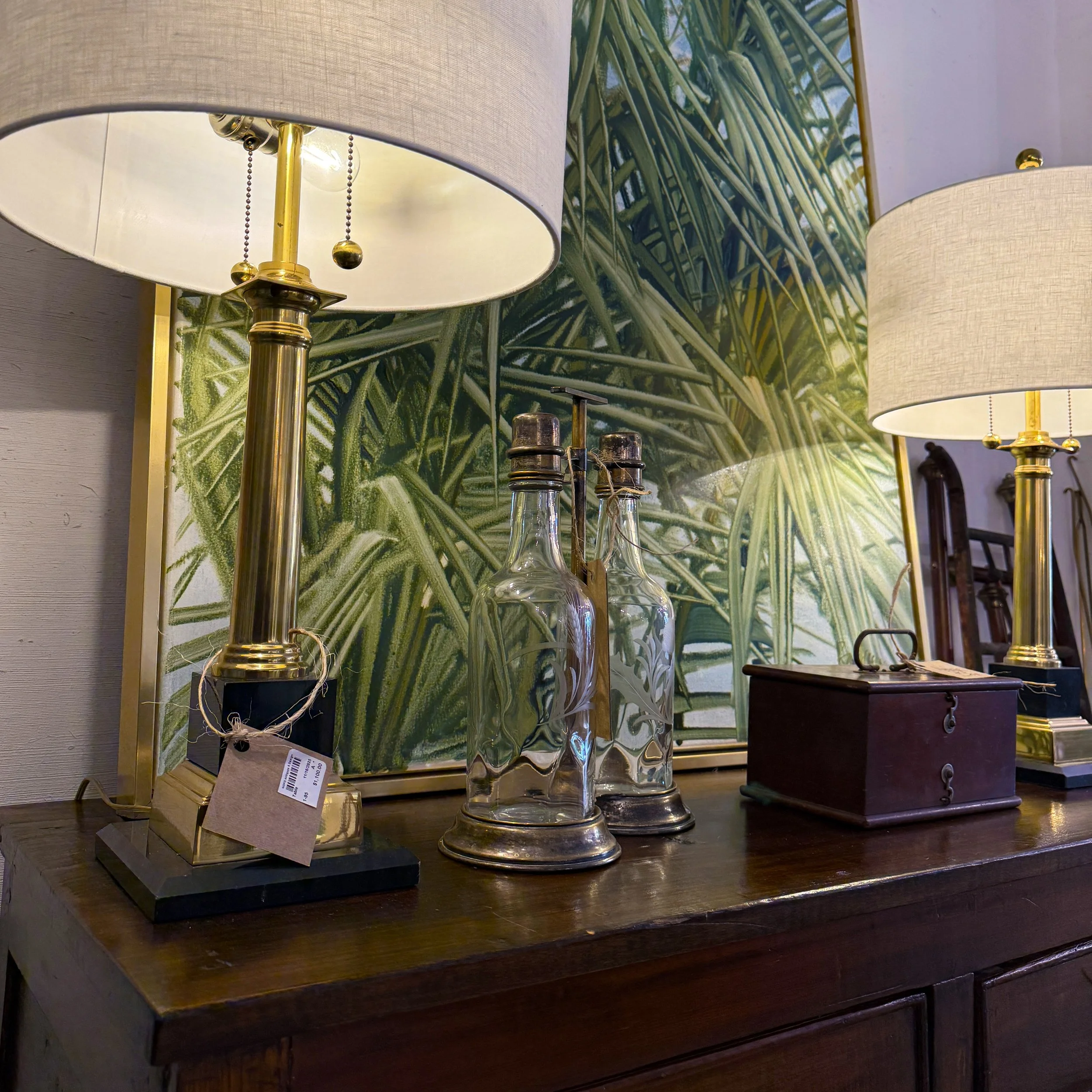 A wooden dresser decorated with two table lamps with brass bases and beige shades, a framed botanical print of green foliage, two vintage glass bottles with metal caps and bases, and a small dark wooden box.