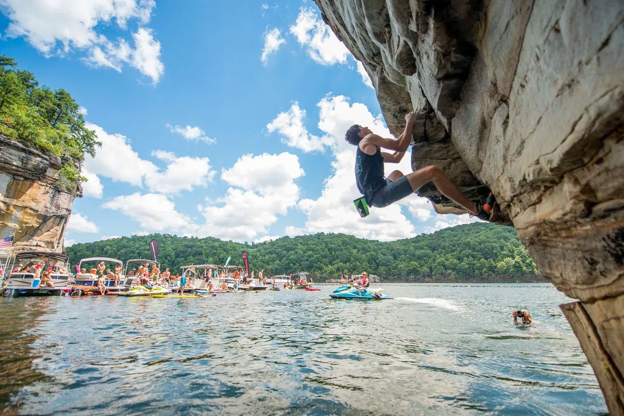 A young man rock climbing on a large overhanging rock beside a body of water on a sunny day with scattered clouds, with boats and people boating in the background.
