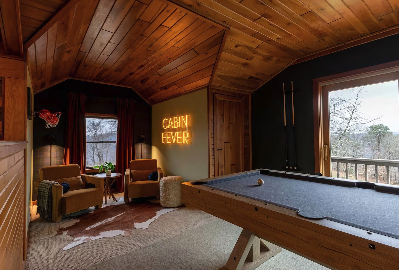 A cozy cabin game room with wood-paneled ceiling and walls, a pool table, a neon sign that reads 'CABIN FEVER,' two armchairs, a small table with a plant, large windows, and a black wall with pool cues hanging.