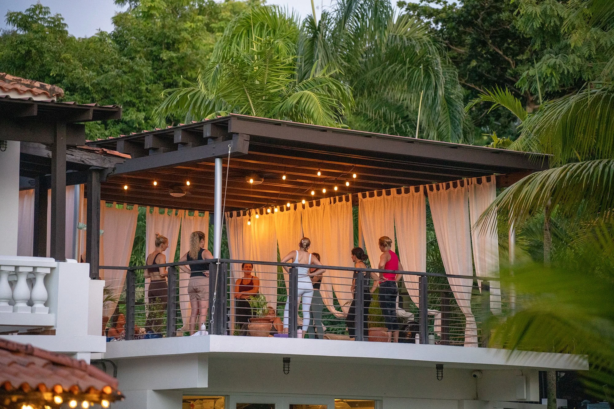 Group of women doing yoga on a covered outdoor terrace at sunset, surrounded by lush tropical trees and decorated with hanging string lights and curtains.