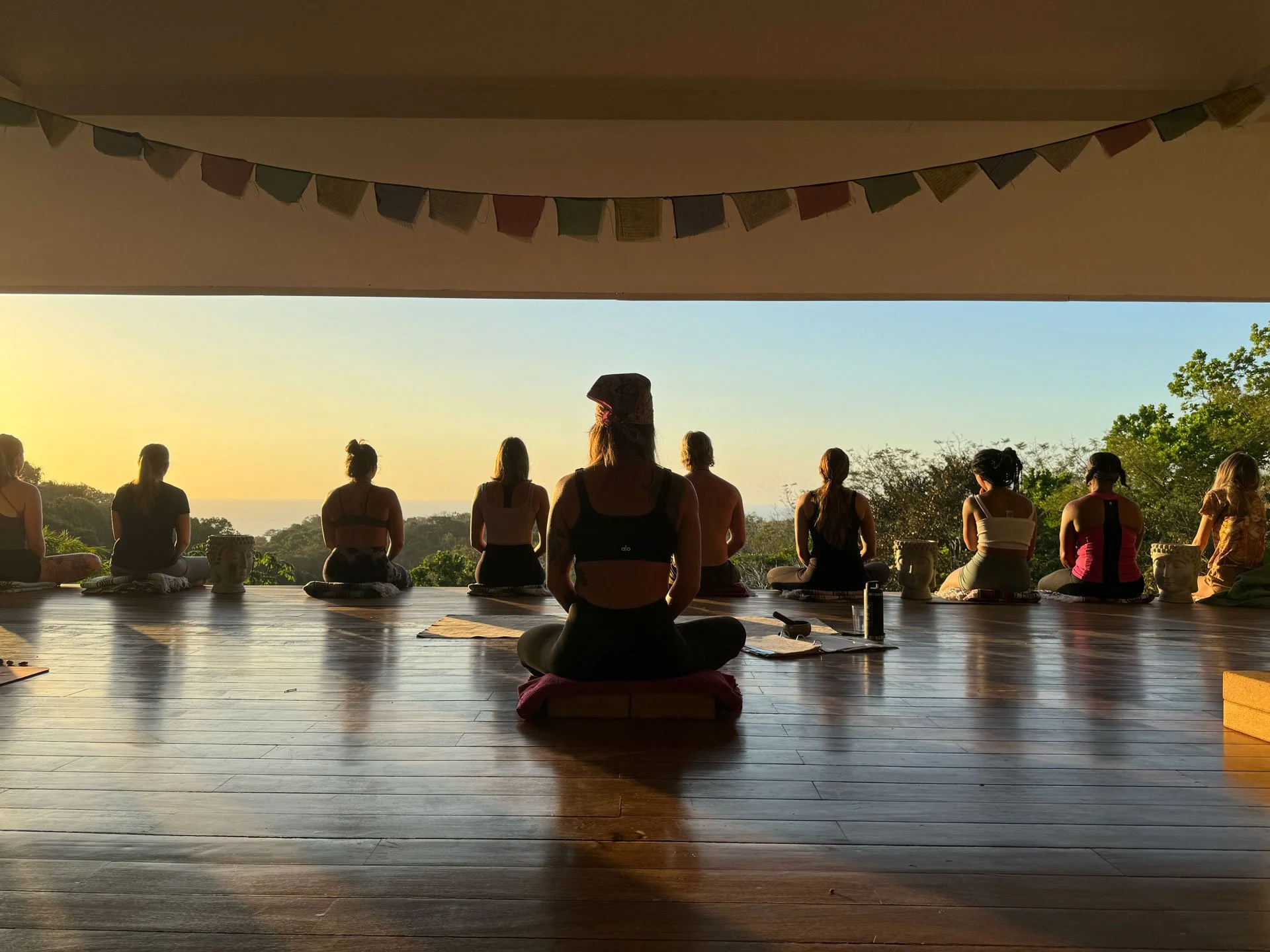 People practicing yoga on a wooden floor in front of an open view of trees and a sunset.