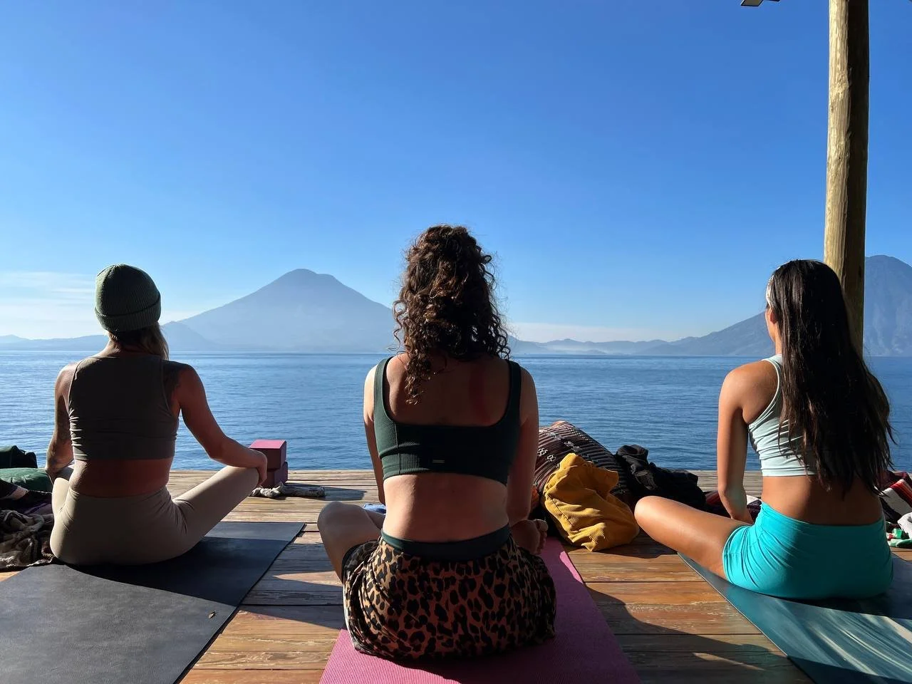 Three women practicing yoga on a wooden deck by a lake, with mountains in the background, during sunny weather.