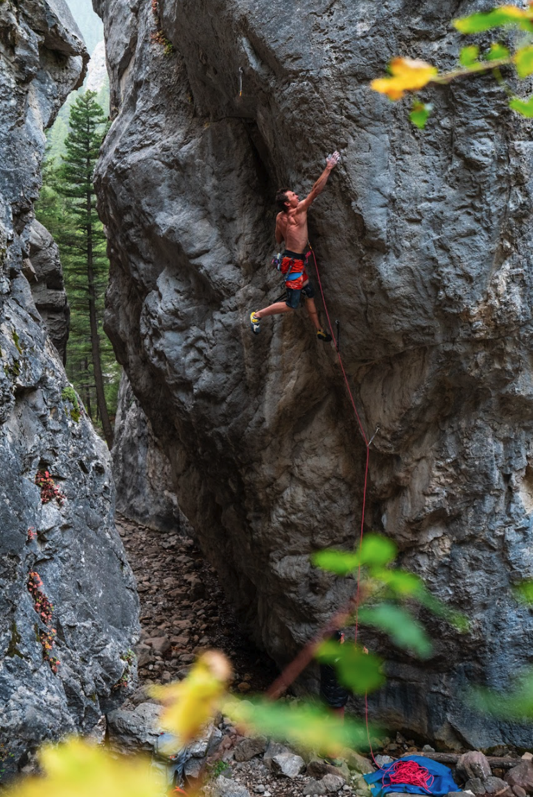 A man climbing a large rock face outdoors, wearing climbing shoes and shorts, with climbing gear and rope, surrounded by trees and rocks.