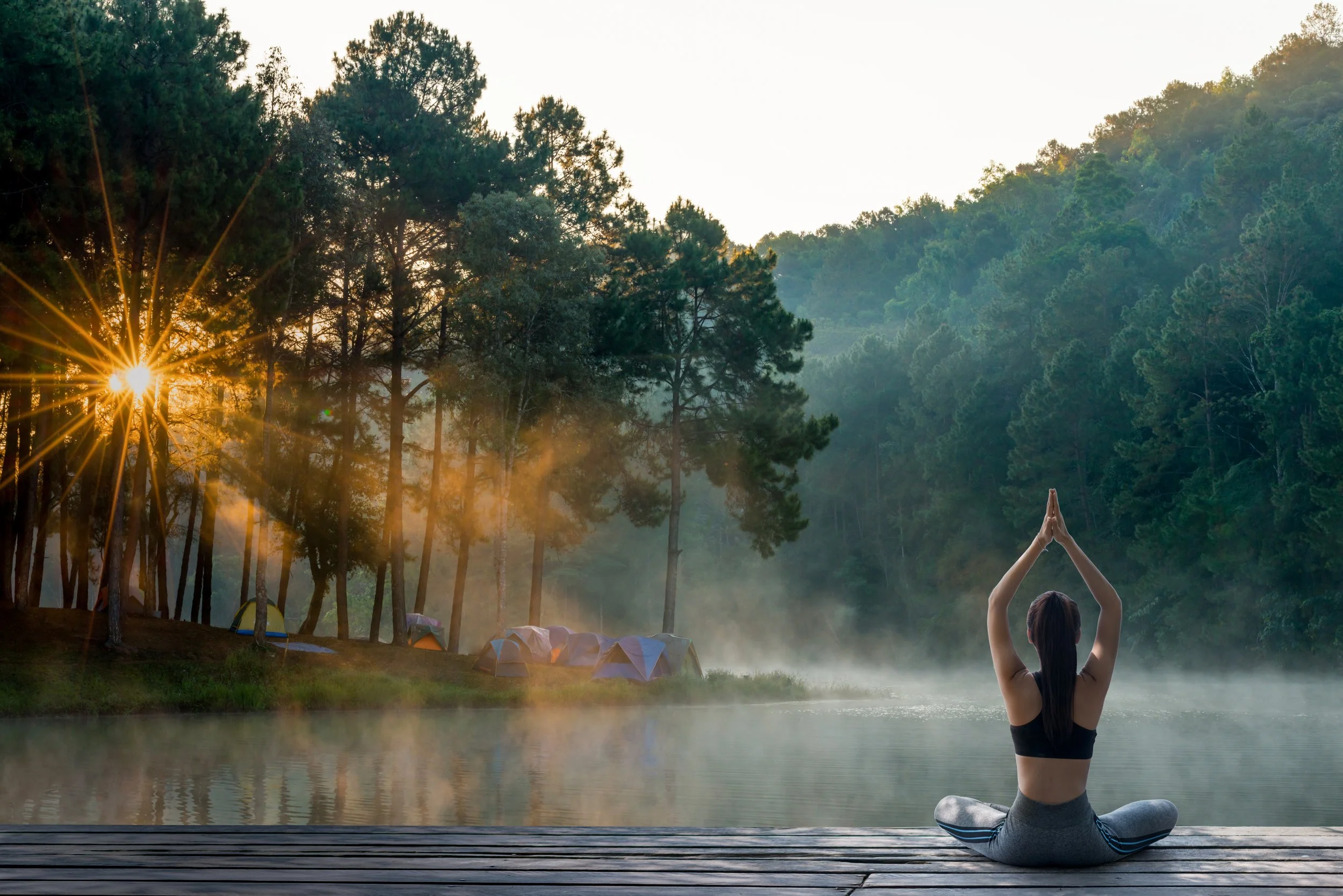 A woman practicing yoga in a seated meditation pose on a wooden dock by a misty lake with tents and trees in the background, during sunrise.