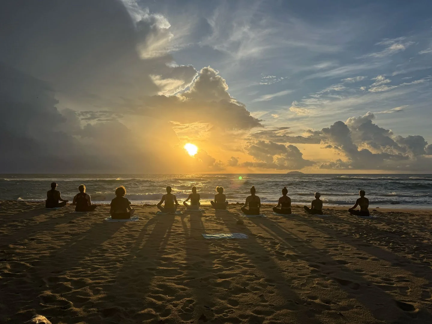 People practicing yoga on the beach during sunset, seated in meditation poses facing the ocean with clouds and the setting sun in the sky.