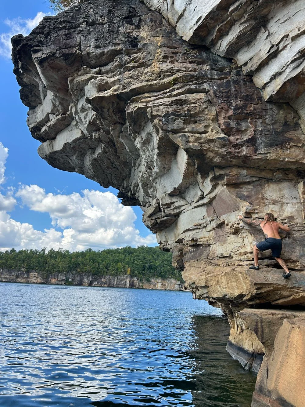 A person rock climbing on a steep cliffside above a body of water, with the shoreline and trees in the background and blue sky with clouds above.