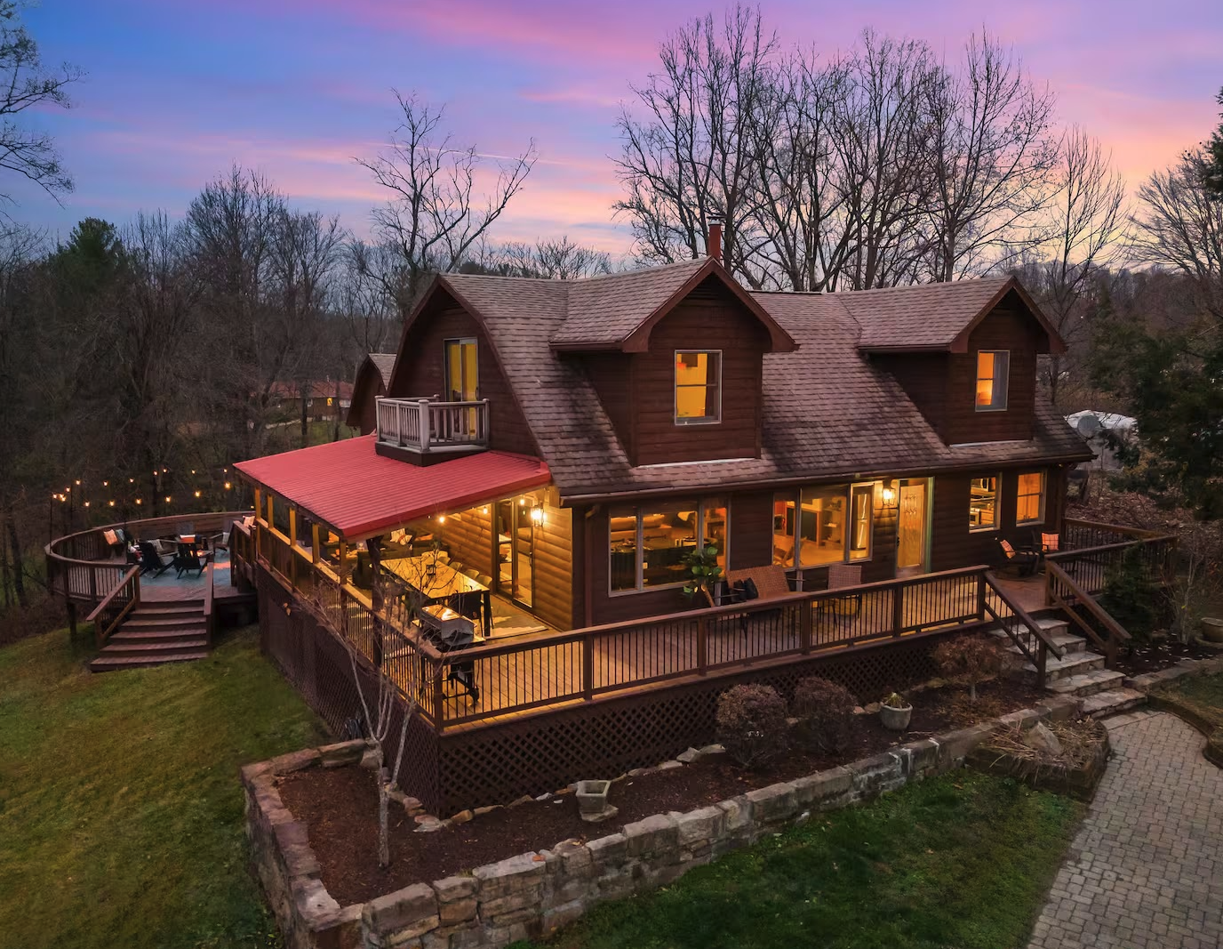 A two-story wooden house with a large elevated deck, lit windows, and string lights, surrounded by trees at sunset.